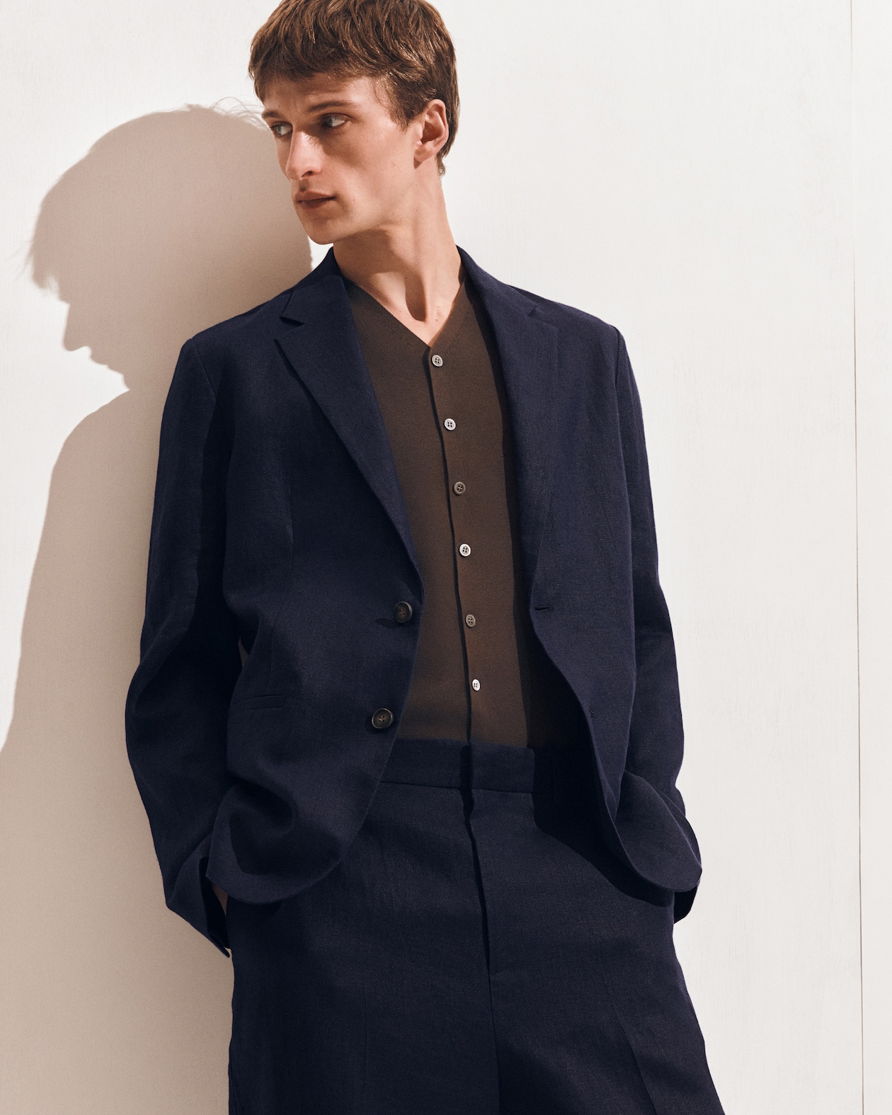Man in a dark suit and brown buttoned shirt, looking to the side against a light background.