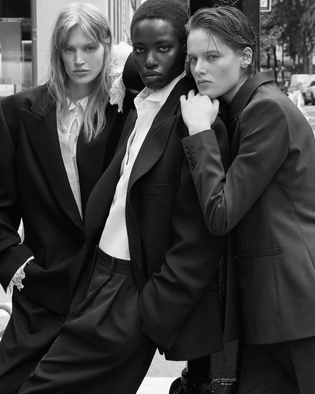 Three models in tailored suits pose confidently outdoors, portraying a sleek and sophisticated fashion style in a black and white photograph.
