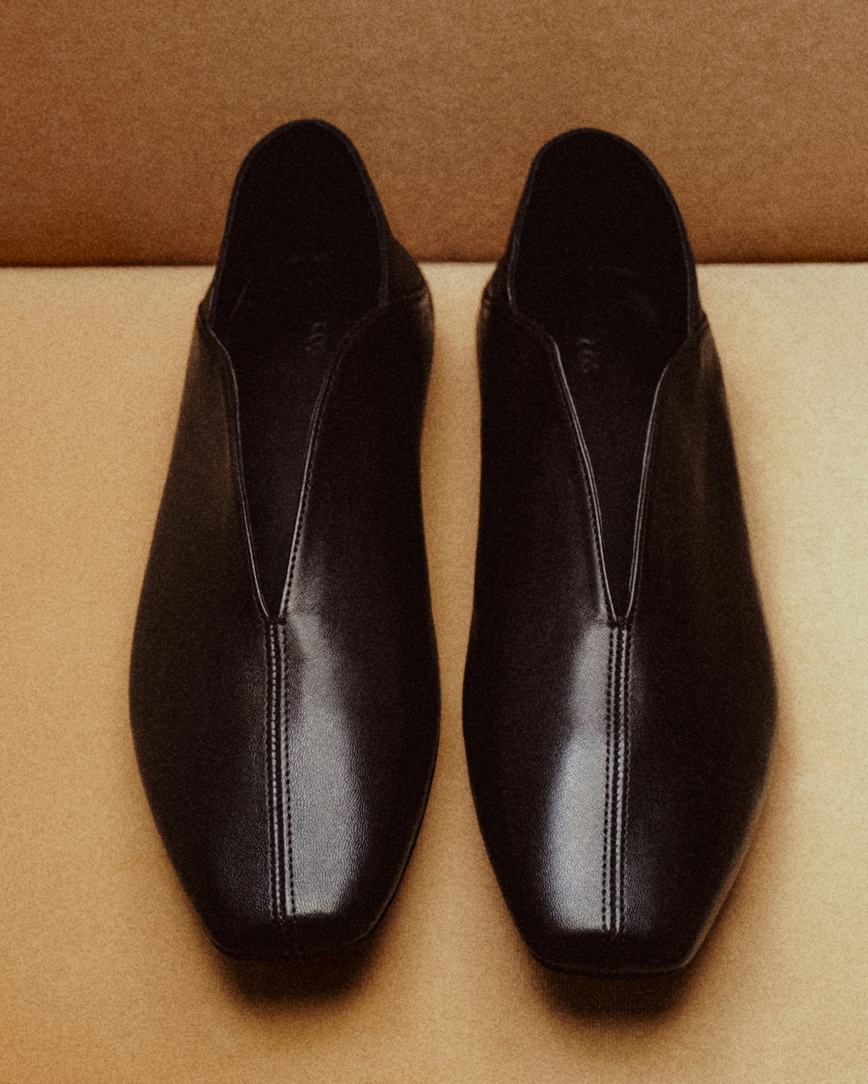 A pair of sleek black leather loafers with square toes and visible stitching, placed side by side on a light brown surface.