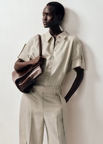 Placeholder for: Person wearing a beige button-up, short-sleeve shirt and matching pants, holding a brown bag, standing against a white background.