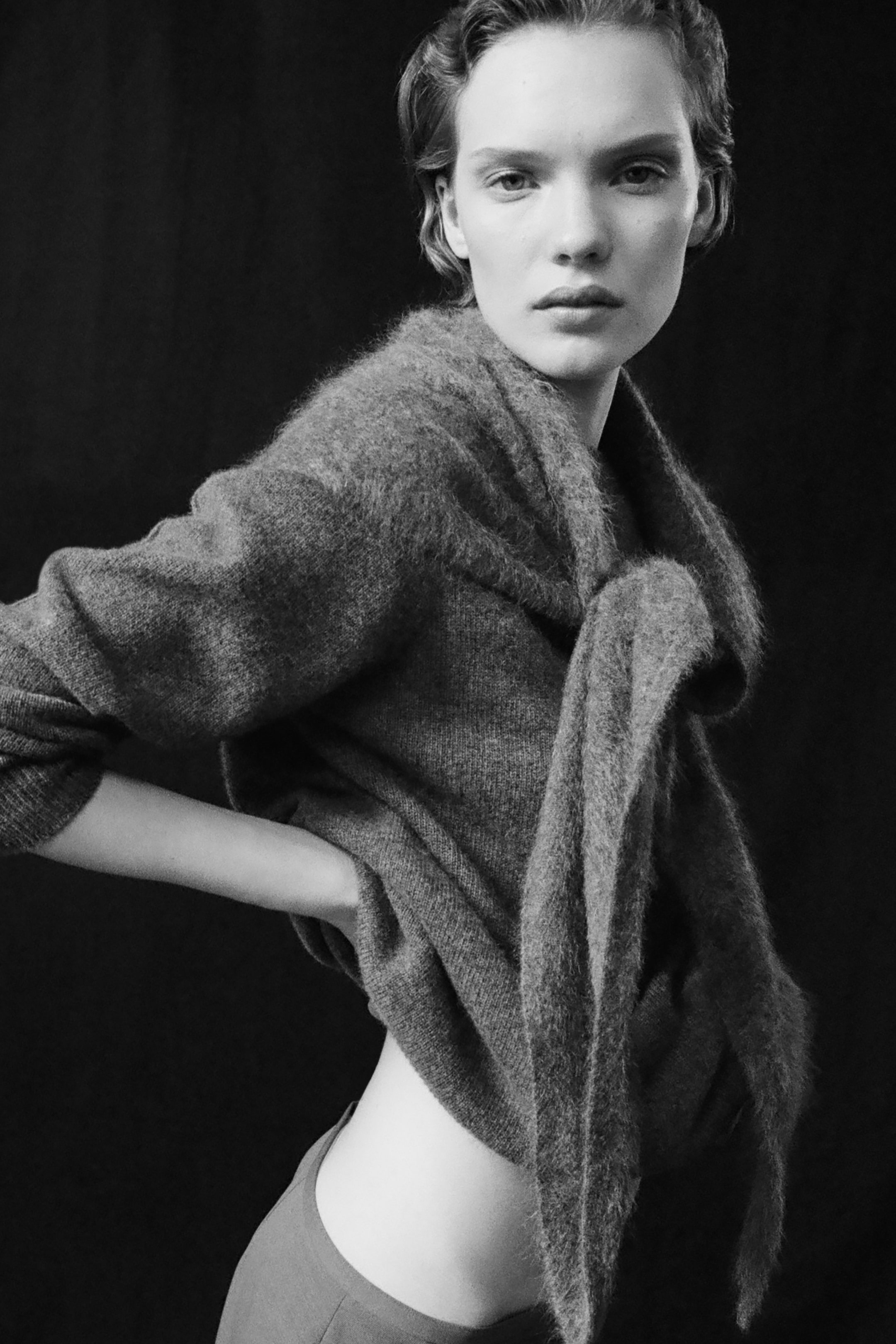 Black and white portrait of a person with short hair, wearing a fuzzy sweater, posing confidently against a dark background.