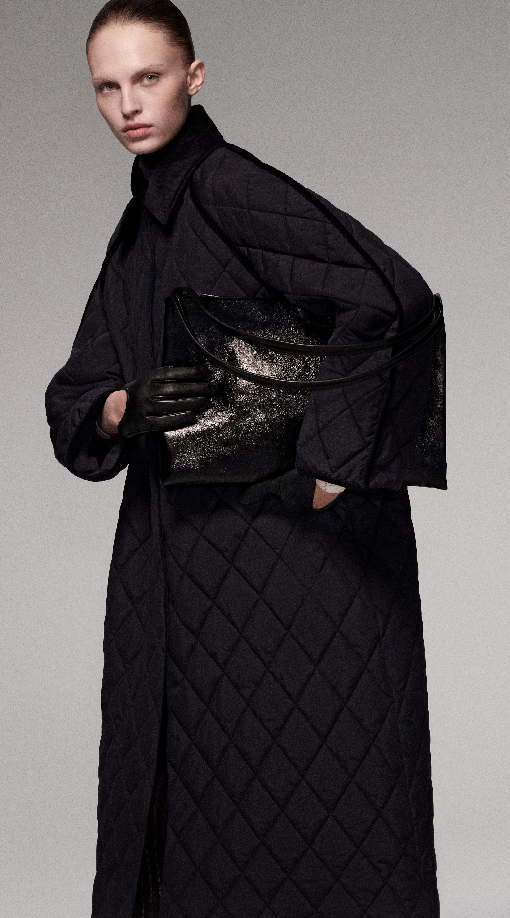 Person in a long, quilted coat, holding a shiny black bag and wearing black gloves, with hair pulled back.