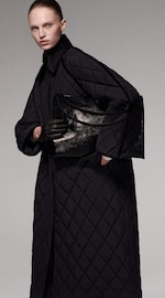 Placeholder for: Person in a long, quilted coat, holding a shiny black bag and wearing black gloves, with hair pulled back.
