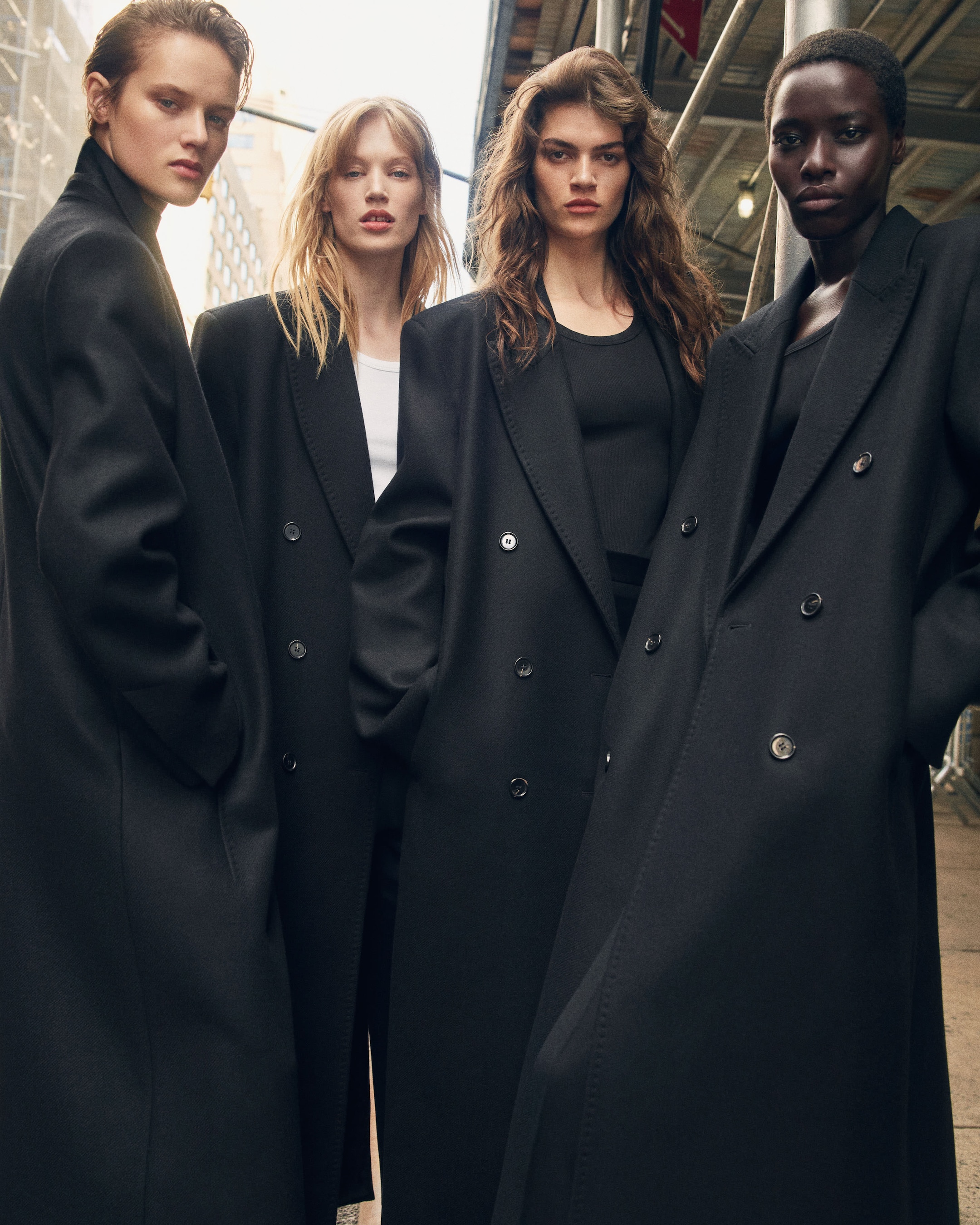 Four people stand in black coats on an urban street, with neutral expressions and a city background.