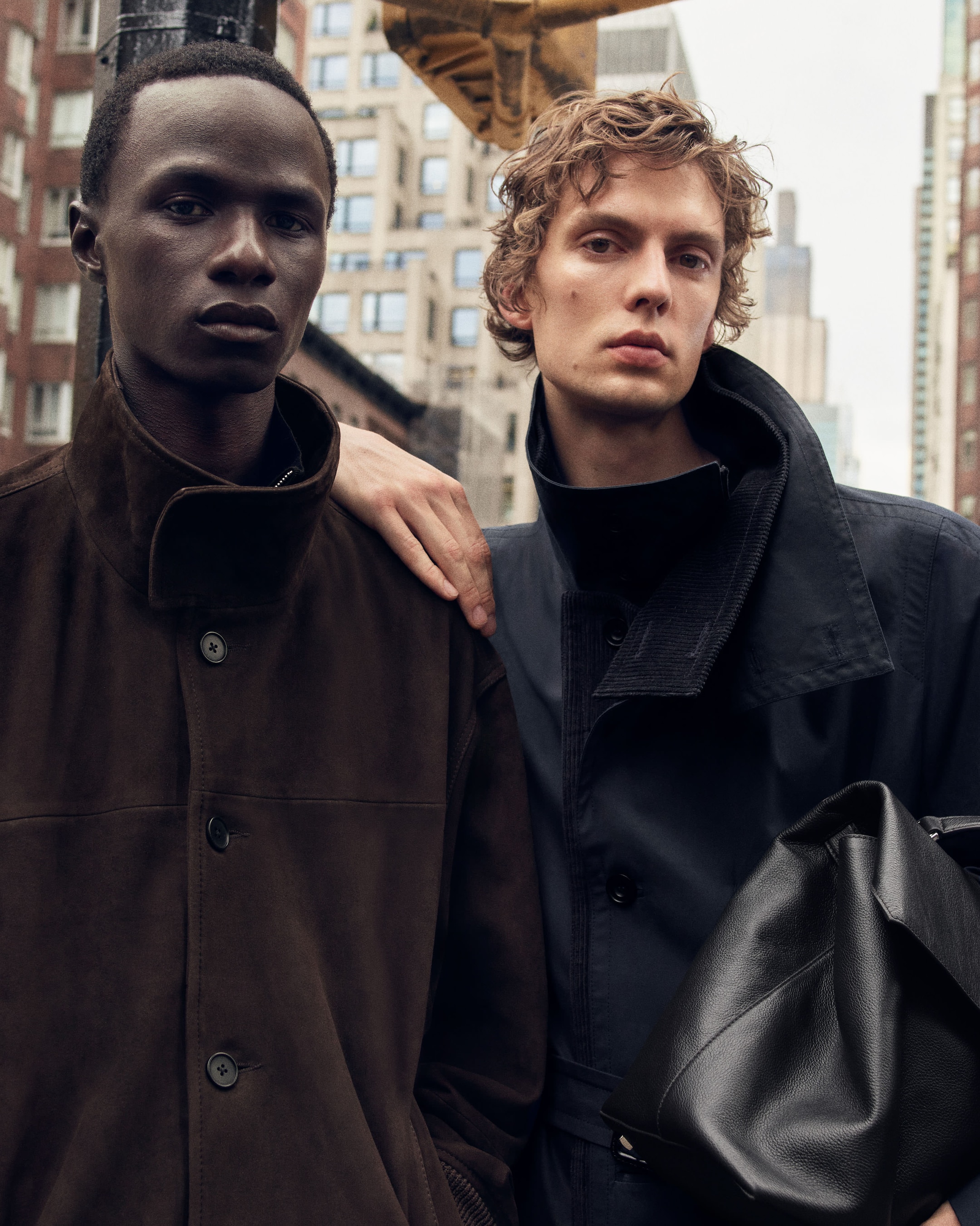 Two individuals in fashionable outerwear stand in an urban setting, one with a hand on the other's shoulder, holding a black leather bag.