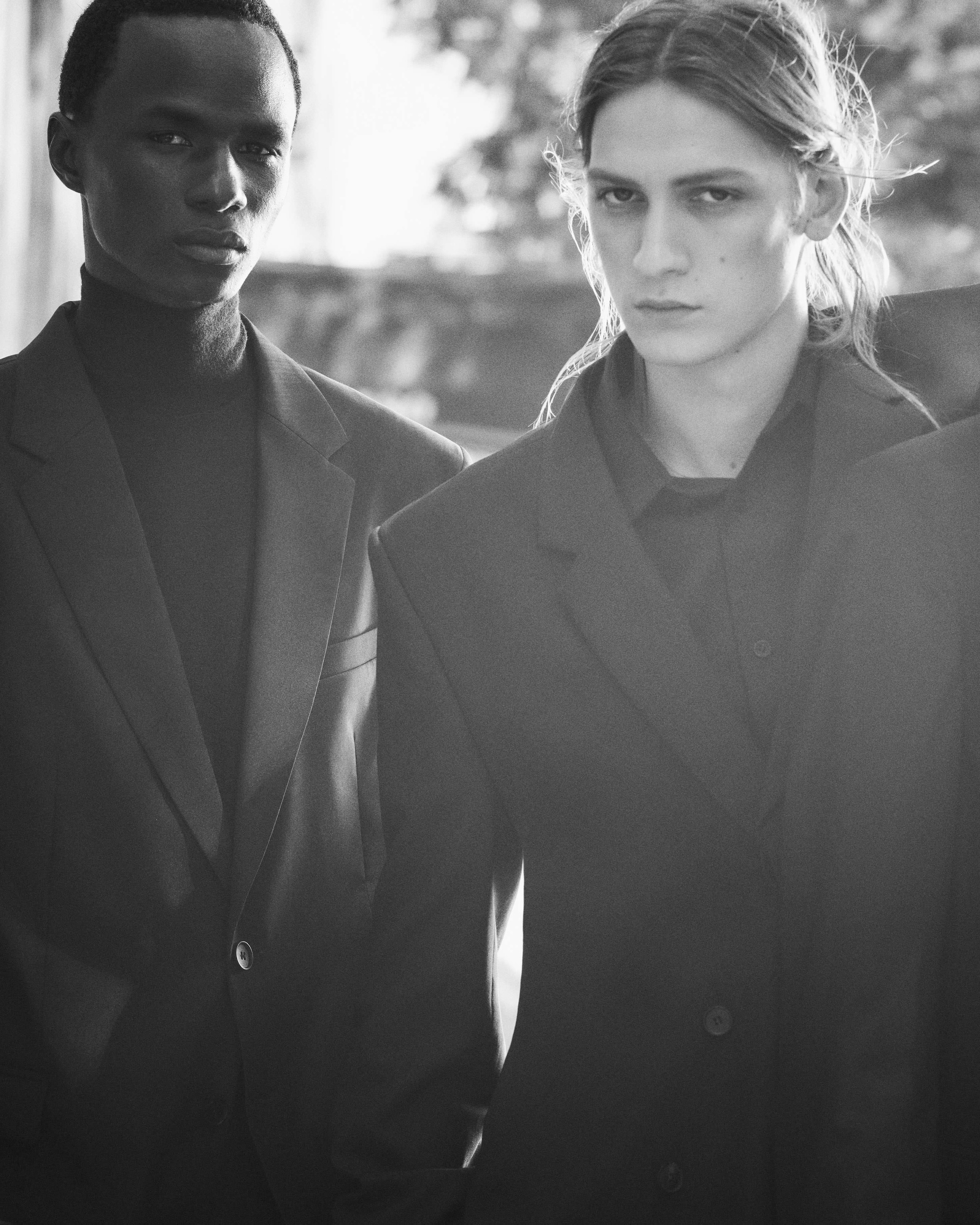 Two individuals in a black-and-white photo are wearing dark suits, standing closely together with serious expressions.
