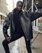 Placeholder for: Person wearing a black leather jacket and leggings poses confidently on an urban street under scaffolding.