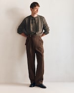 Placeholder for: Man in a casual cardigan and high-waisted trousers leans against a white wall, hands in pockets, looking to the side.