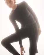 Placeholder for: Person in a fitted, long-sleeve, gray outfit poses elegantly against a soft, light background. They are leaning forward with one leg raised.