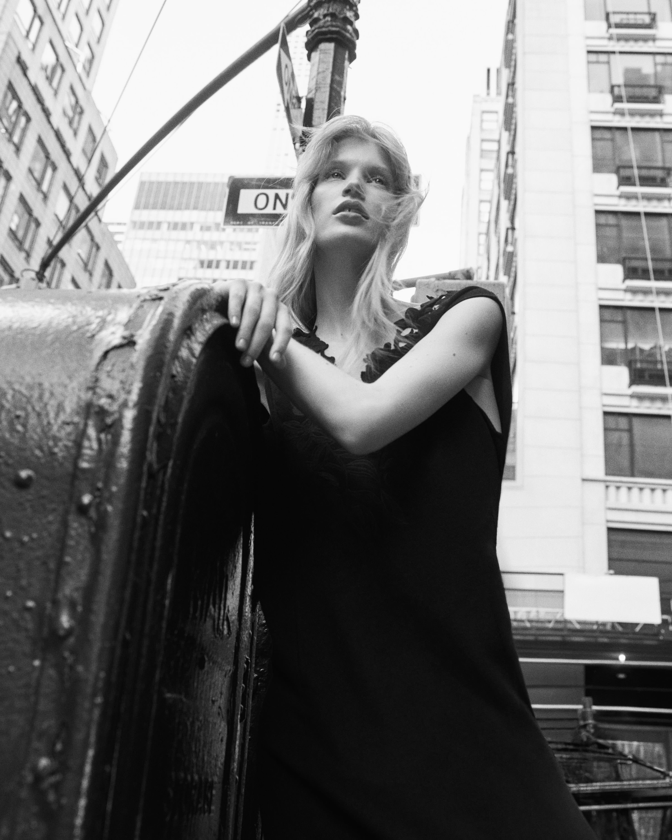 Woman in a black dress stands on a city street corner, leaning against a gritty metal surface, with buildings and a one-way sign in the background.
