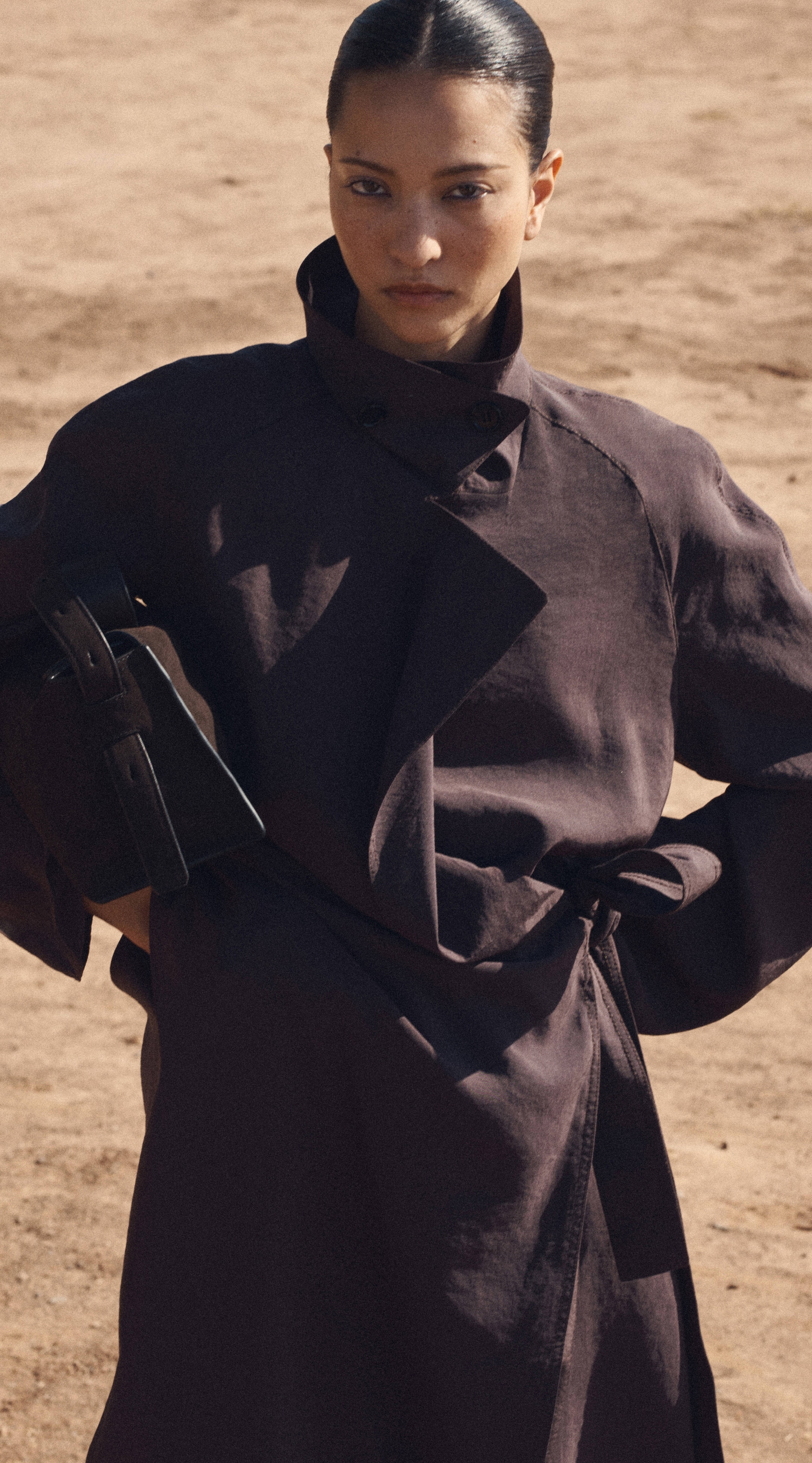 Person in a high-collared, dark coat stands confidently on a sandy terrain, holding a brown bag.