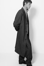 Placeholder for: Person in a long, dark overcoat and trousers, wearing a light turtleneck, walking against a plain white background.