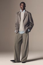 Placeholder for: Person wearing grey blazer, cable-knit sweater, blue shirt, wide-legged trousers, and black shoes stands against a neutral background.