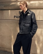 Placeholder for: Person with long hair wearing a black leather jacket and pants, standing against a stone wall with "5 PA" partially visible.