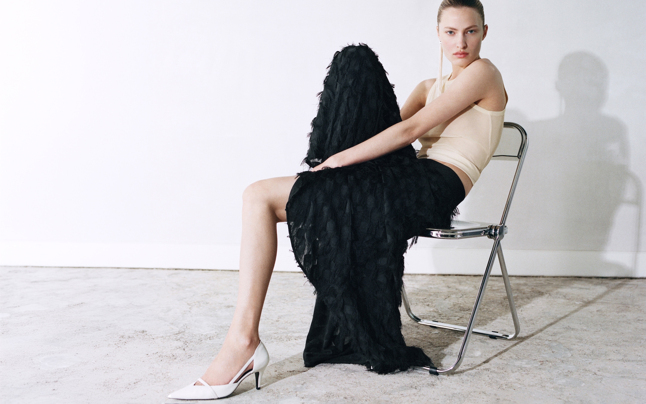 Person seated on a metal chair, wearing a beige top, black textured skirt, and white heels, against a plain white background.