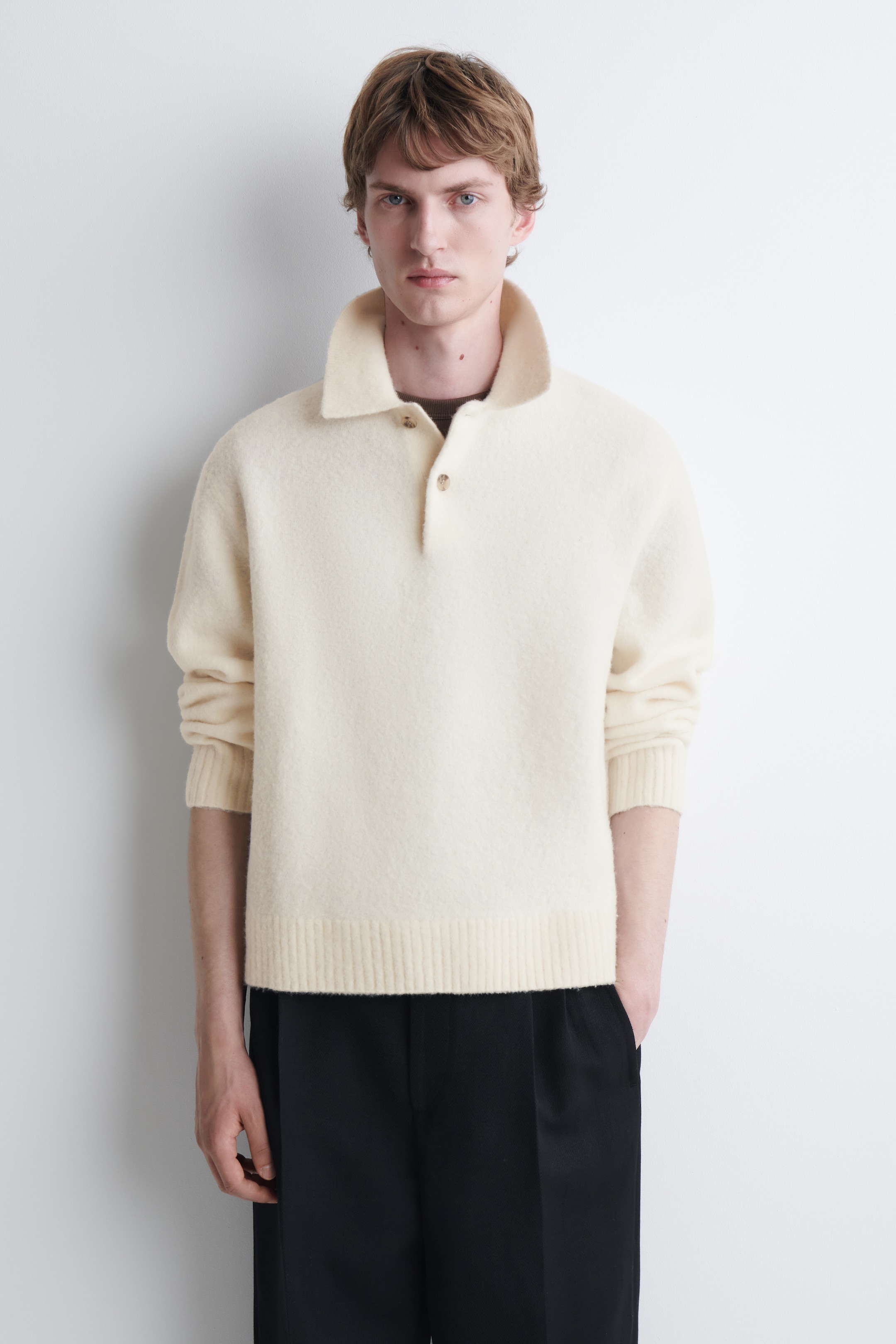 A person with short hair wearing a cream-colored woolen sweater with a high collar and black pants against a plain background.