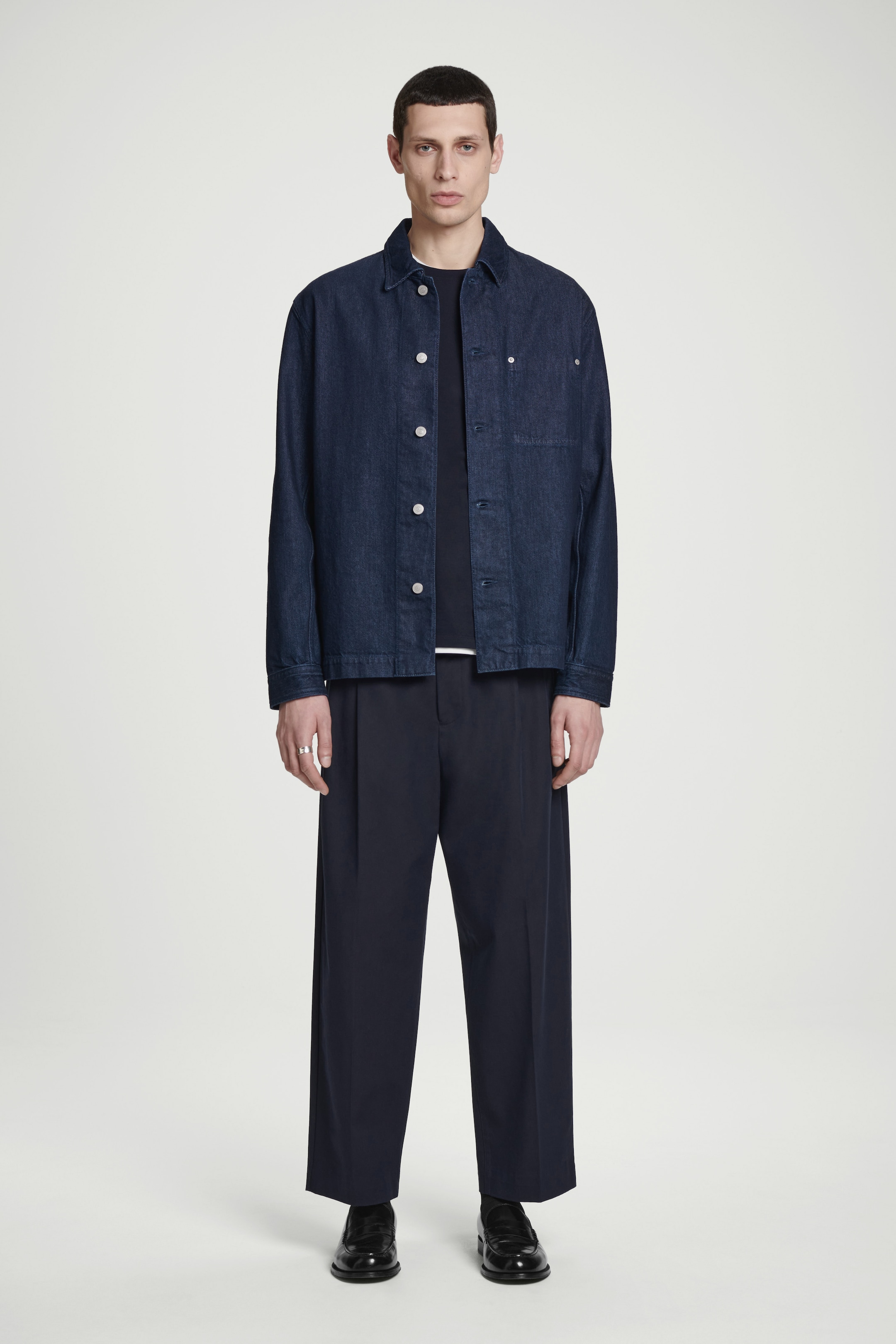 A person wearing a dark denim jacket, black shirt, wide-leg pants, and black shoes stands against a plain white background.