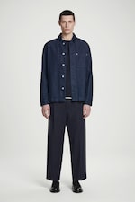 Placeholder for: A person wearing a dark denim jacket, black shirt, wide-leg pants, and black shoes stands against a plain white background.