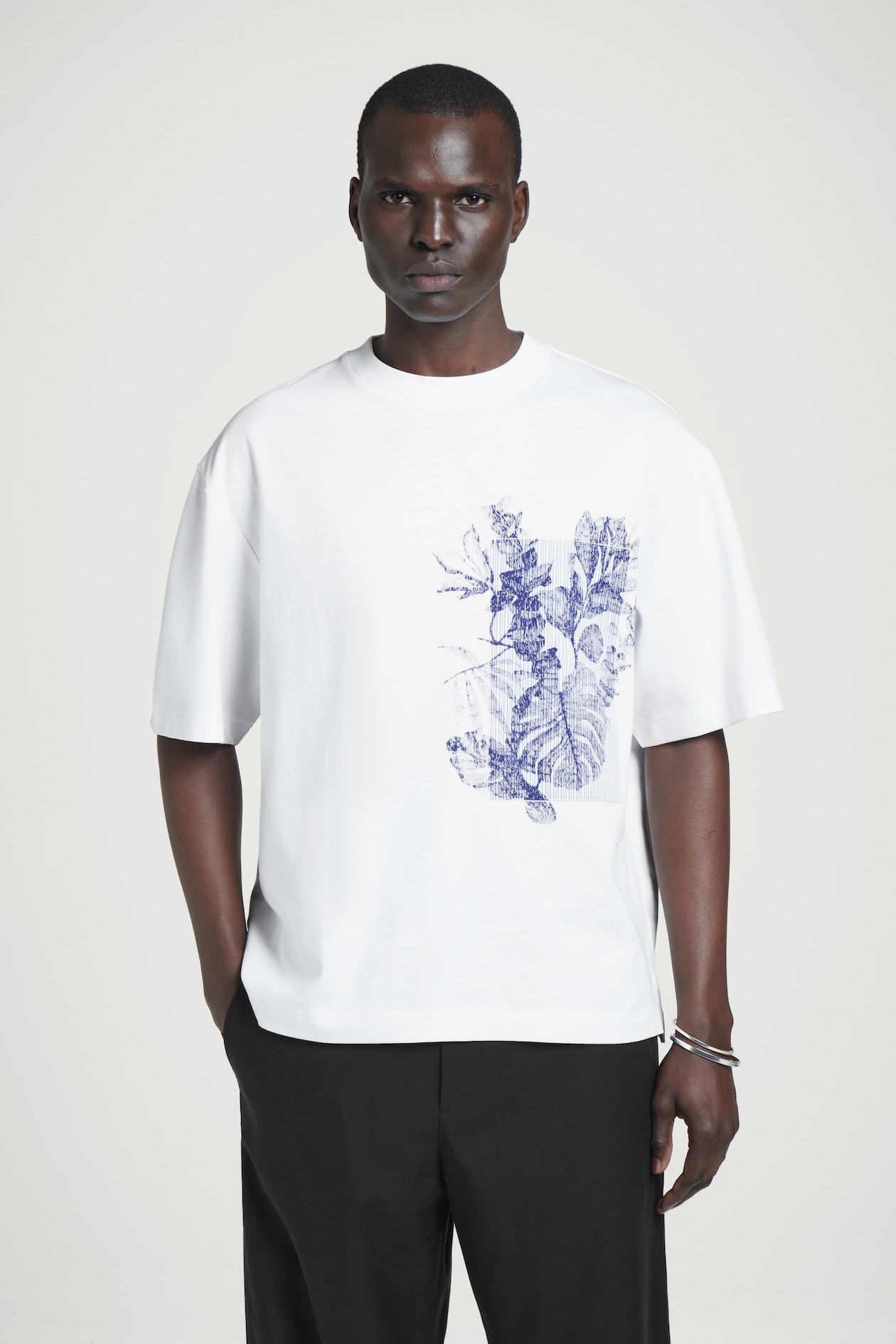 Man wearing a white T-shirt with a blue abstract design and black pants, standing against a plain background.