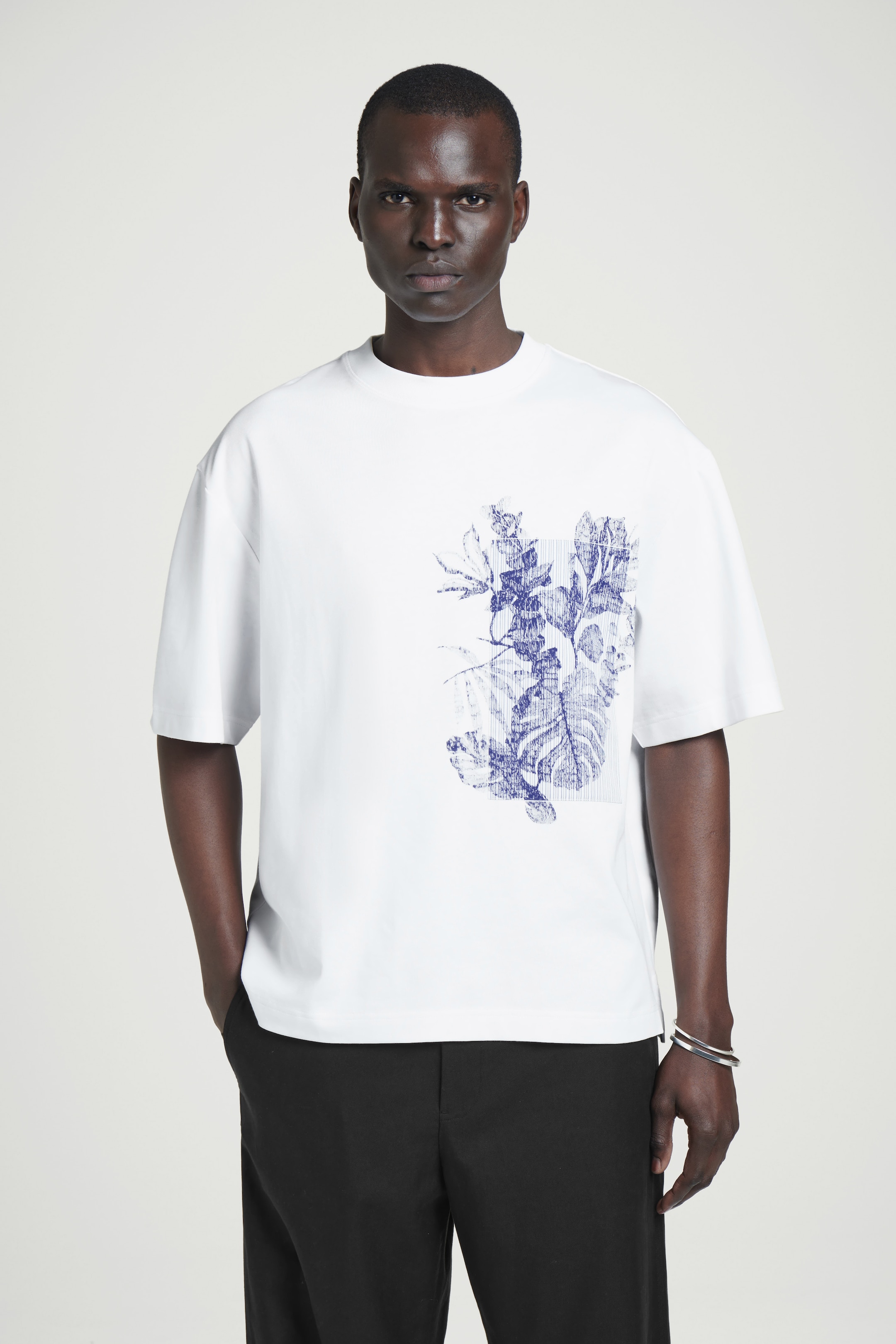Man wearing a white T-shirt with a blue abstract design and black pants, standing against a plain background.