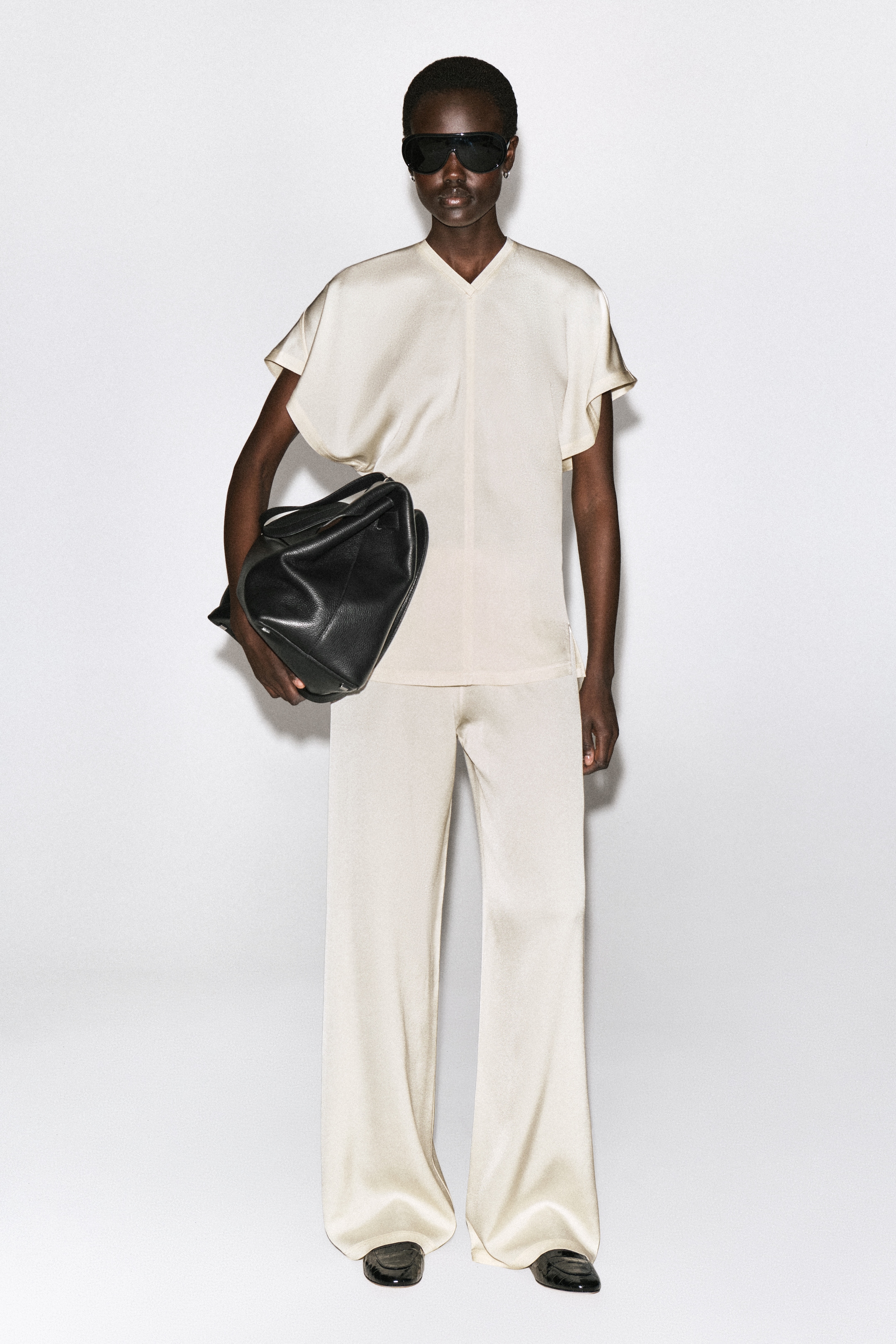 A person wearing beige silk outfit and sunglasses, holding a black leather bag, stands against a white background.