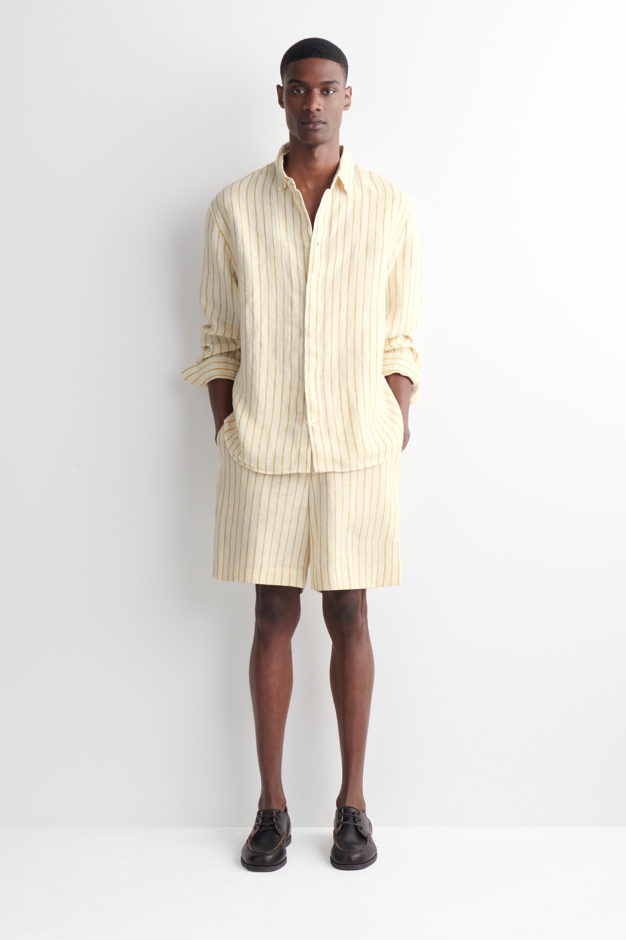 Man wearing a beige and yellow striped shirt with matching shorts, hands in pockets, black shoes, stands against a white background.