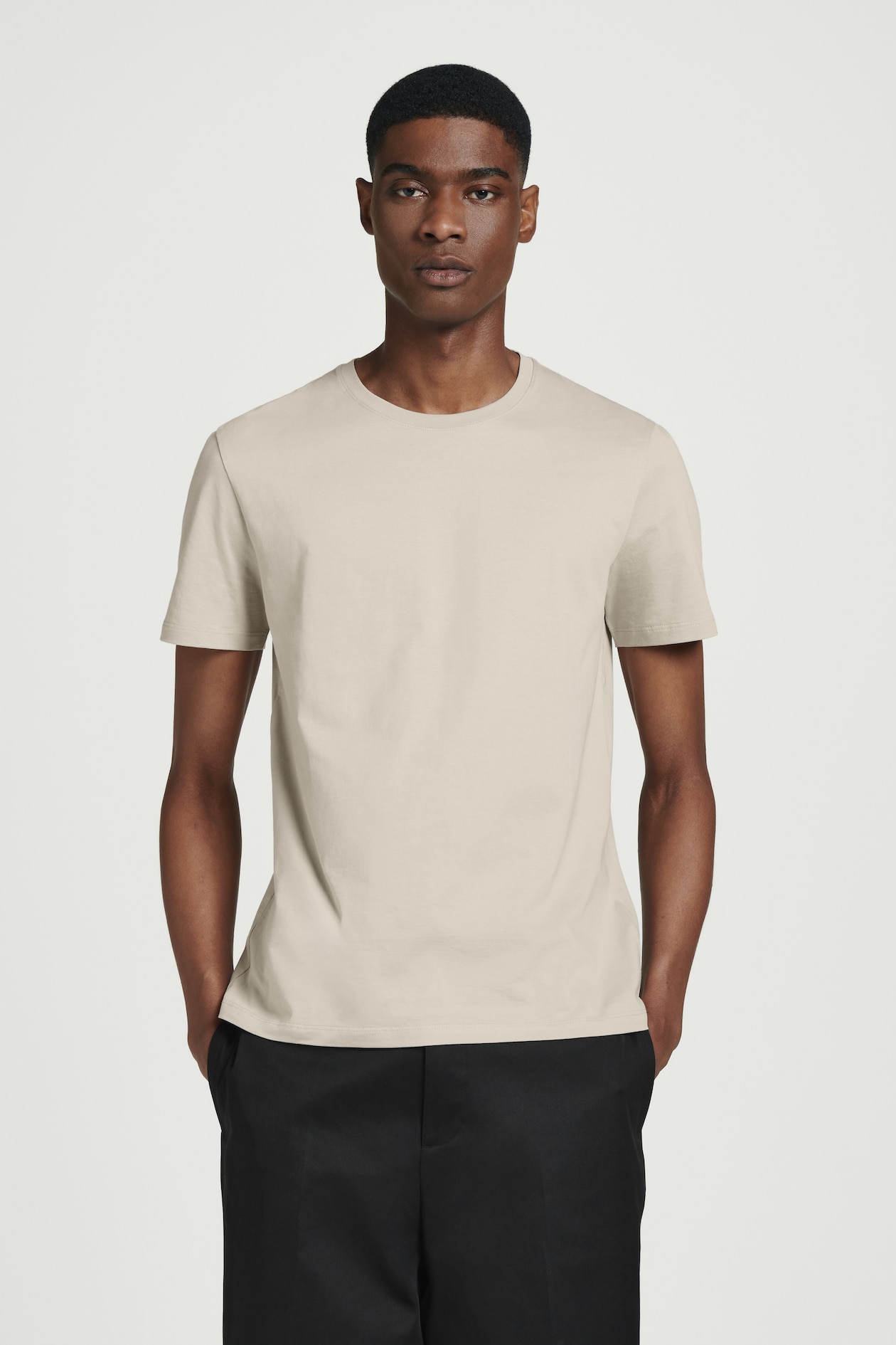 Person wearing a beige t-shirt and black pants, standing with hands in pockets against a plain white background.