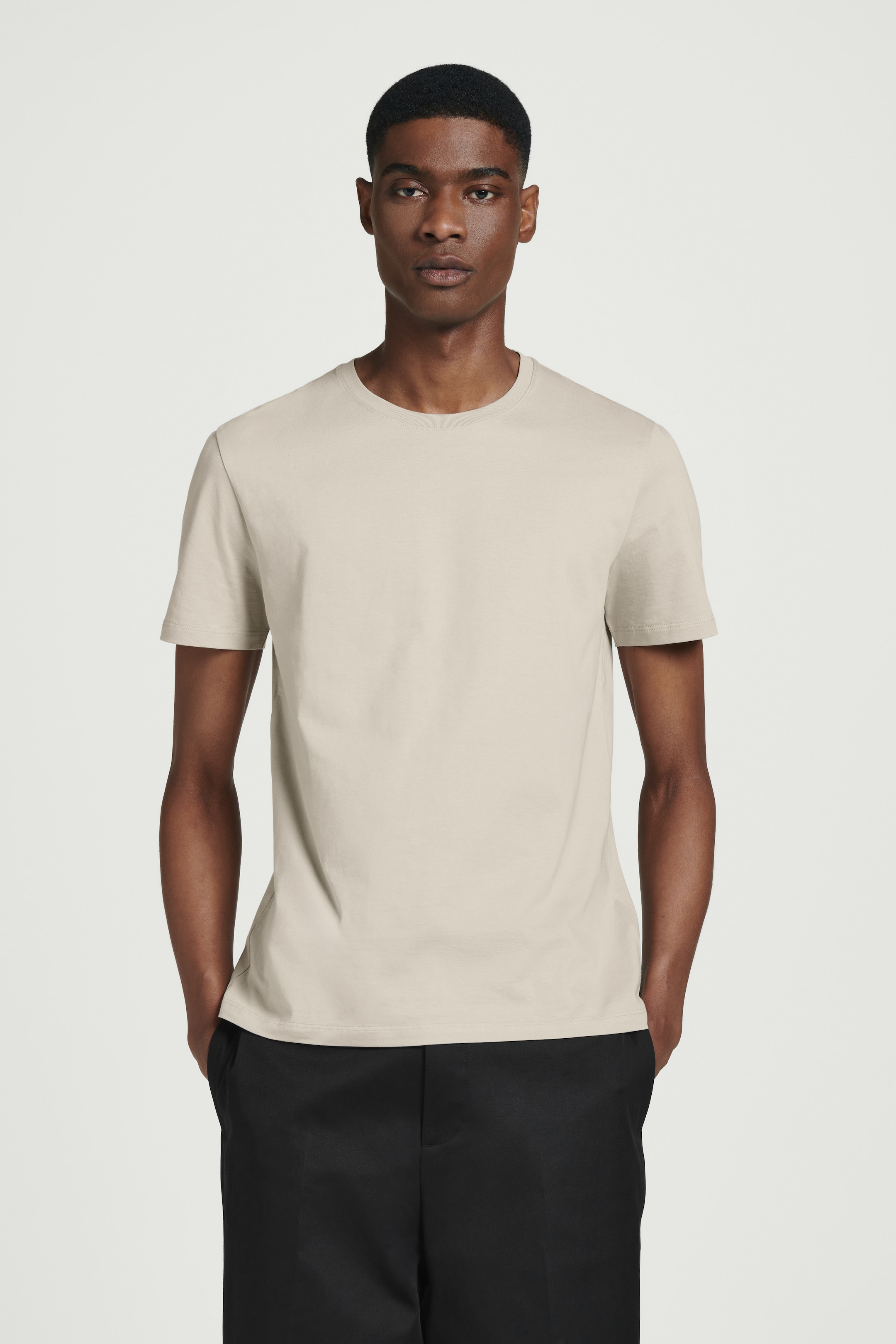 Person wearing a beige t-shirt and black pants, standing with hands in pockets against a plain white background.