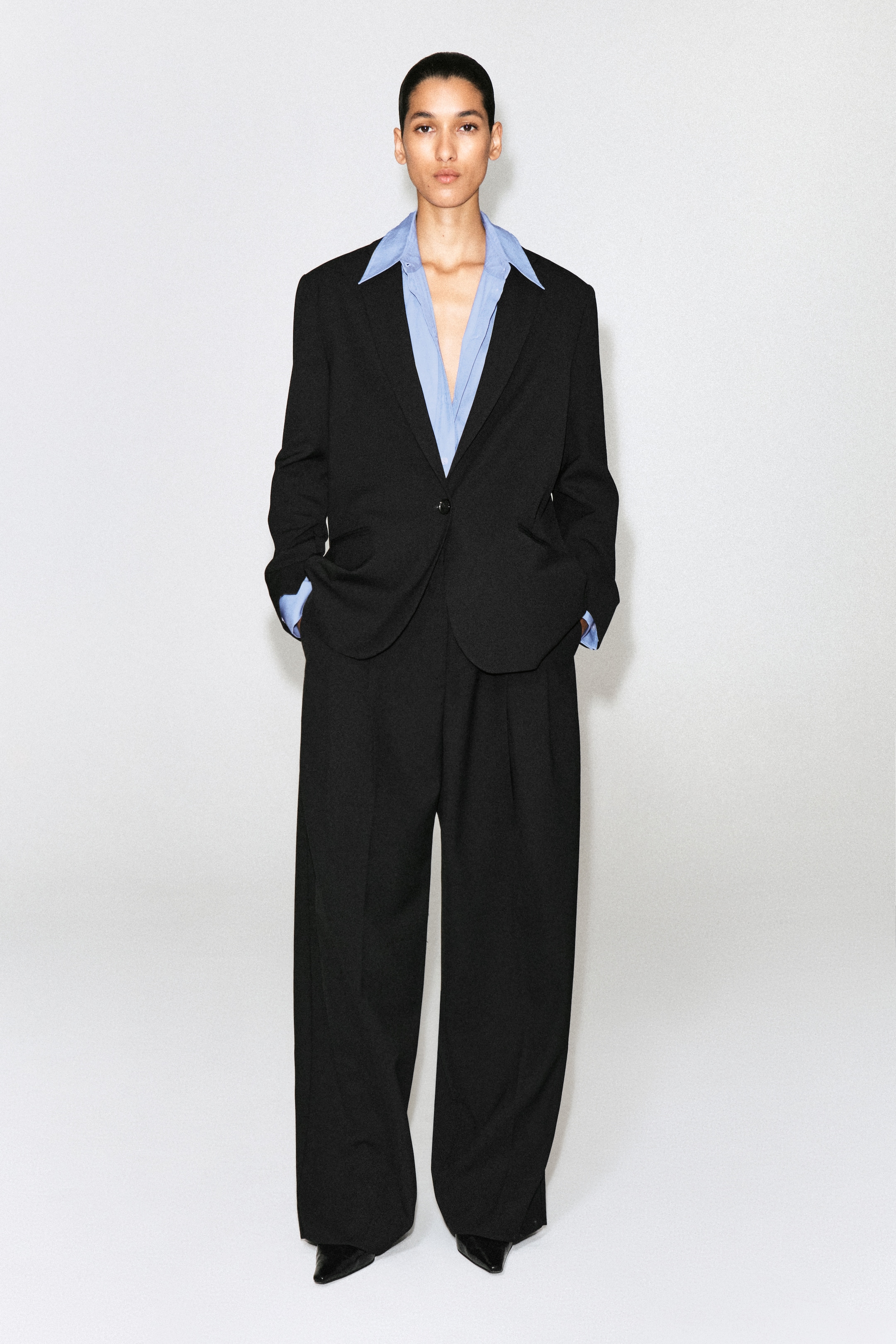 Person wearing an oversized black suit with a light blue dress shirt, standing confidently against a plain background.