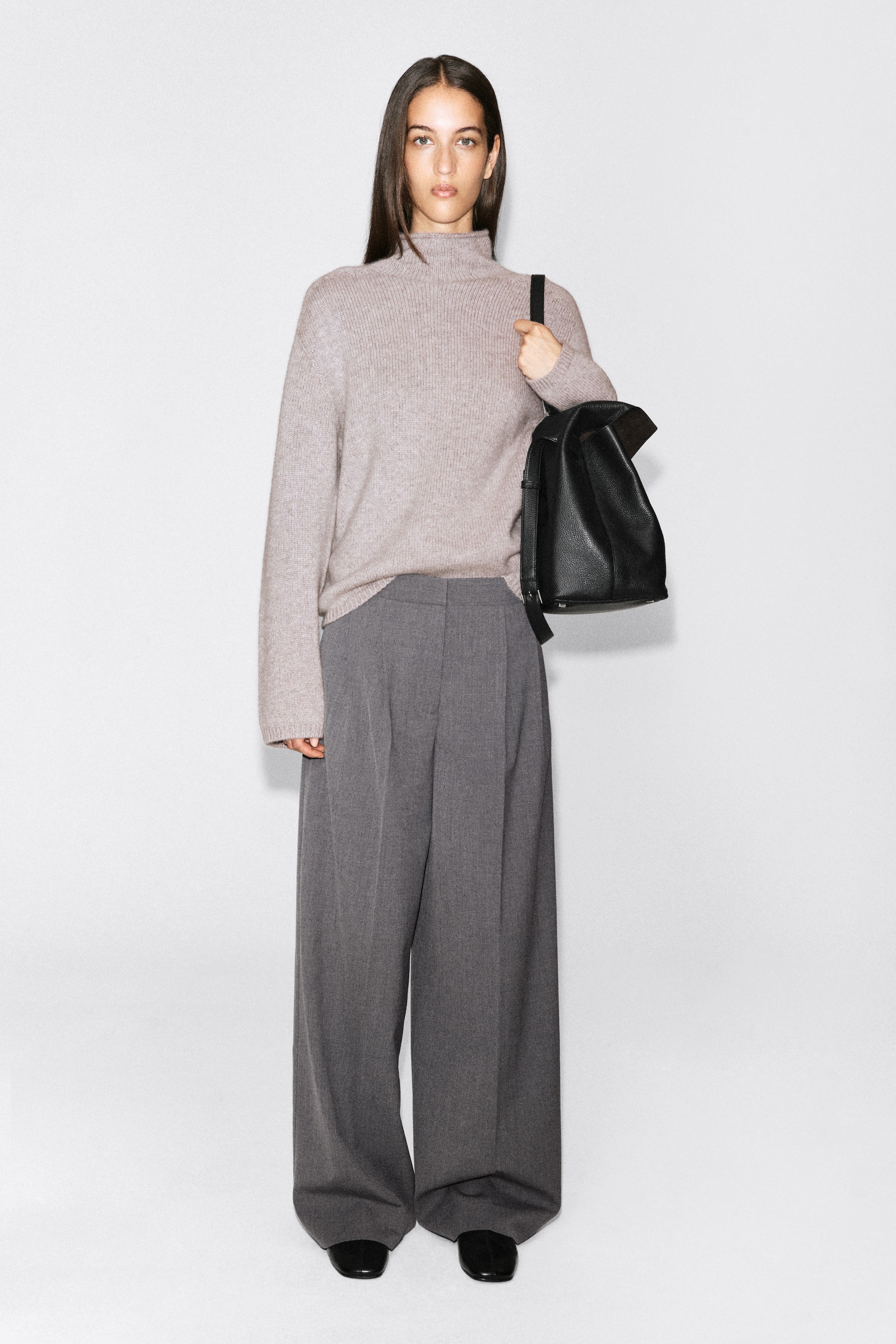 A person stands wearing a beige turtleneck sweater, grey wide-leg trousers, and holding a black leather bag.