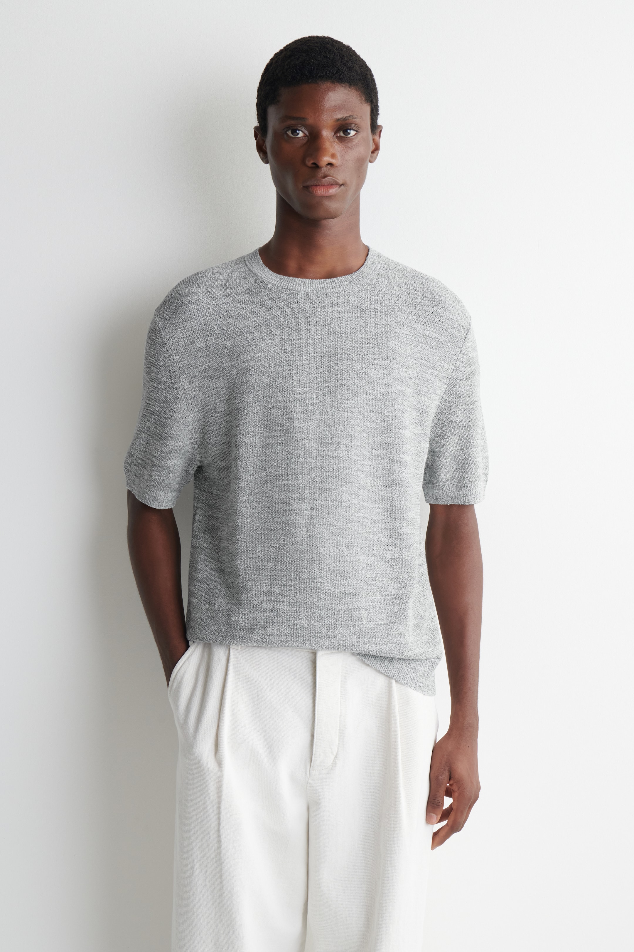 Young man in a gray short-sleeve sweater and white pants, standing against a white background with hands in pockets.