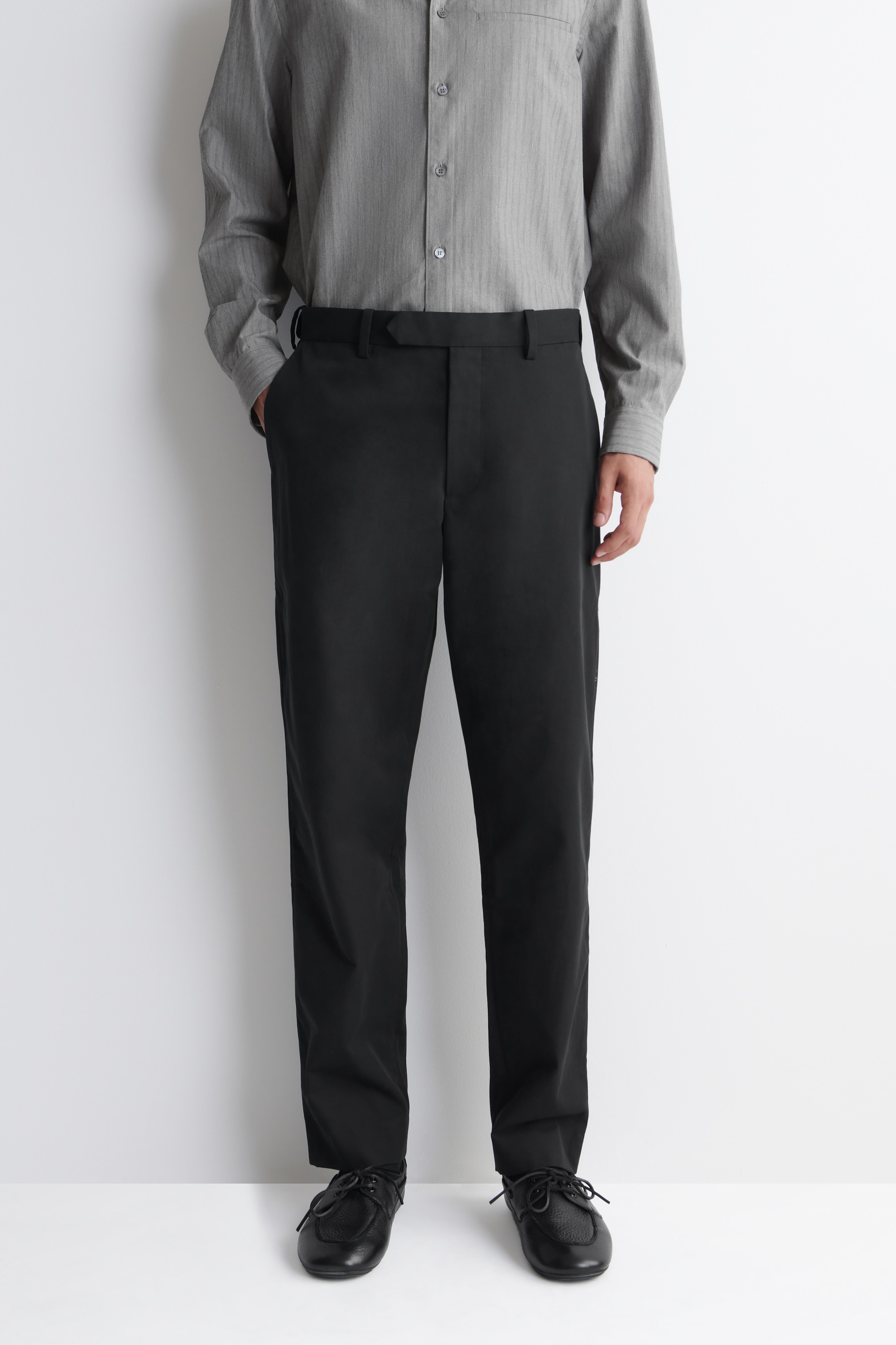 Person wearing a grey long-sleeve shirt and black pants paired with black leather shoes, standing against a white background.