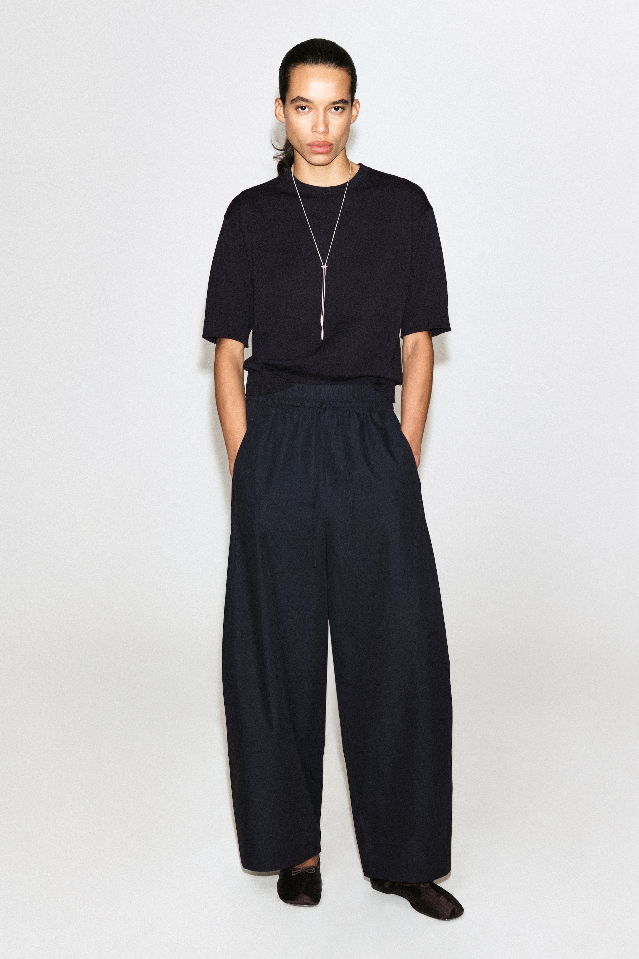 Person wearing a black shirt, wide-leg pants, and a silver necklace, standing against a plain white background.