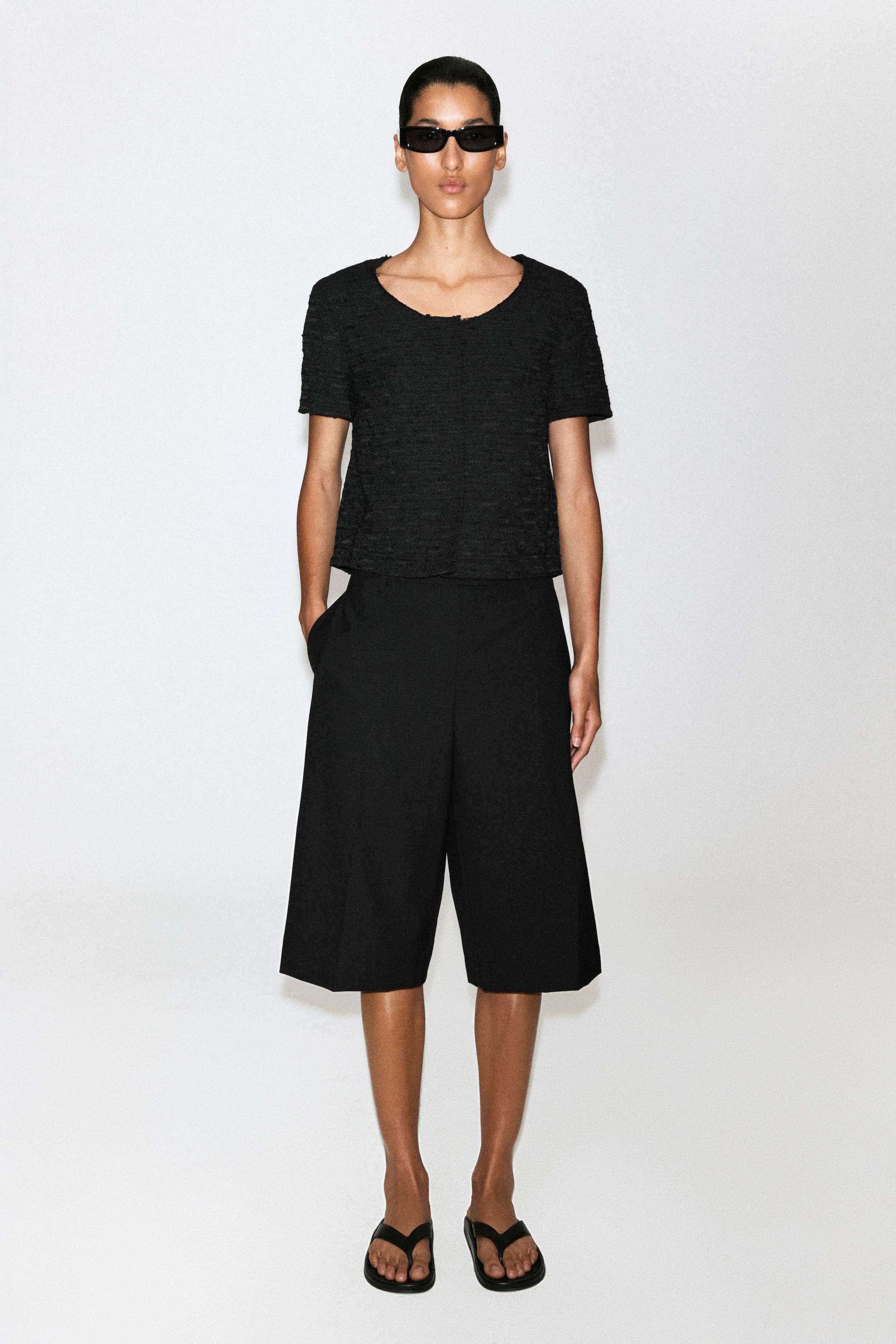 A person wearing black sunglasses, a short-sleeve top, and culottes with hands in pockets, standing against a plain white background.