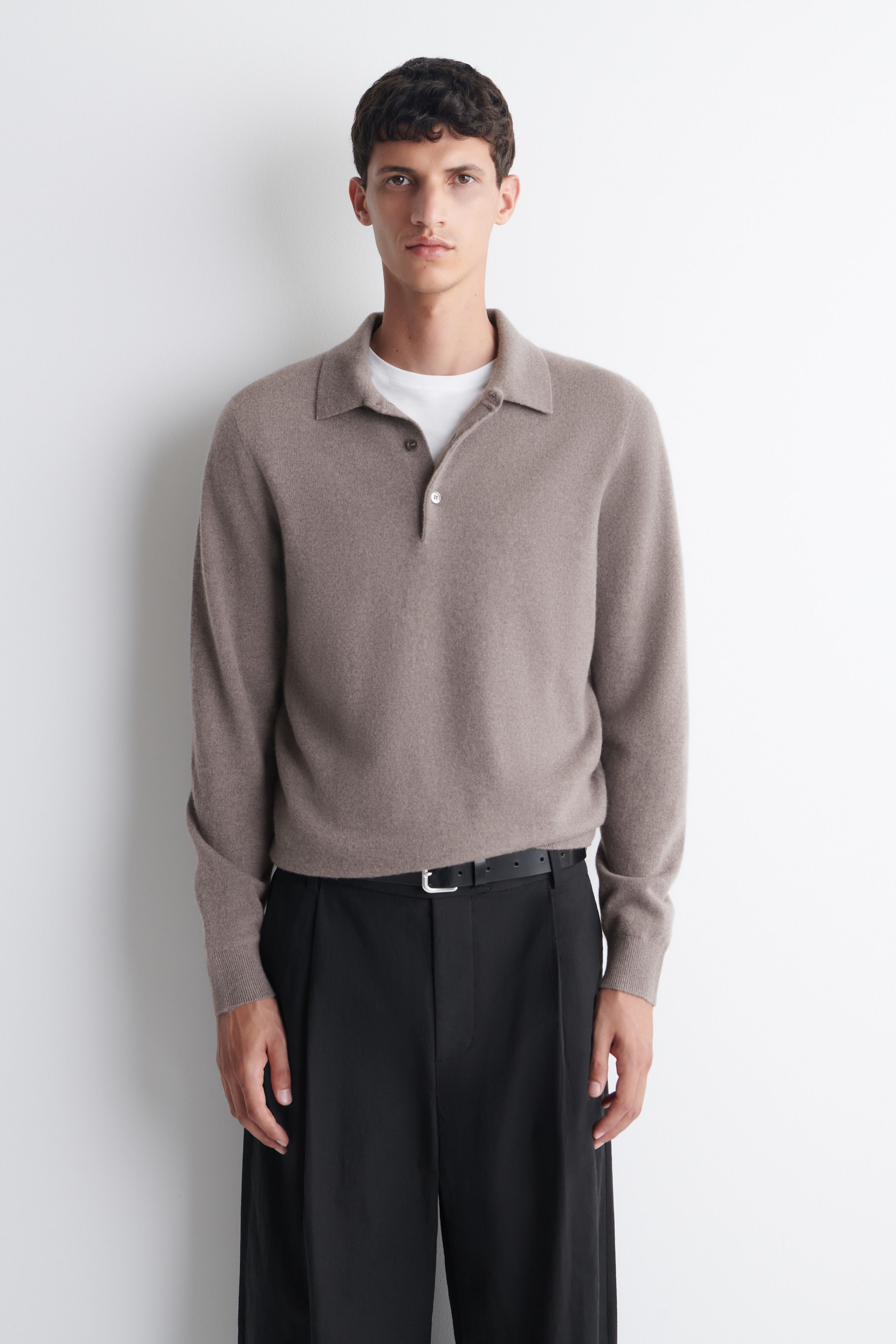A person wearing a taupe long-sleeve polo over a white shirt, paired with black trousers and a belt, standing against a white background.