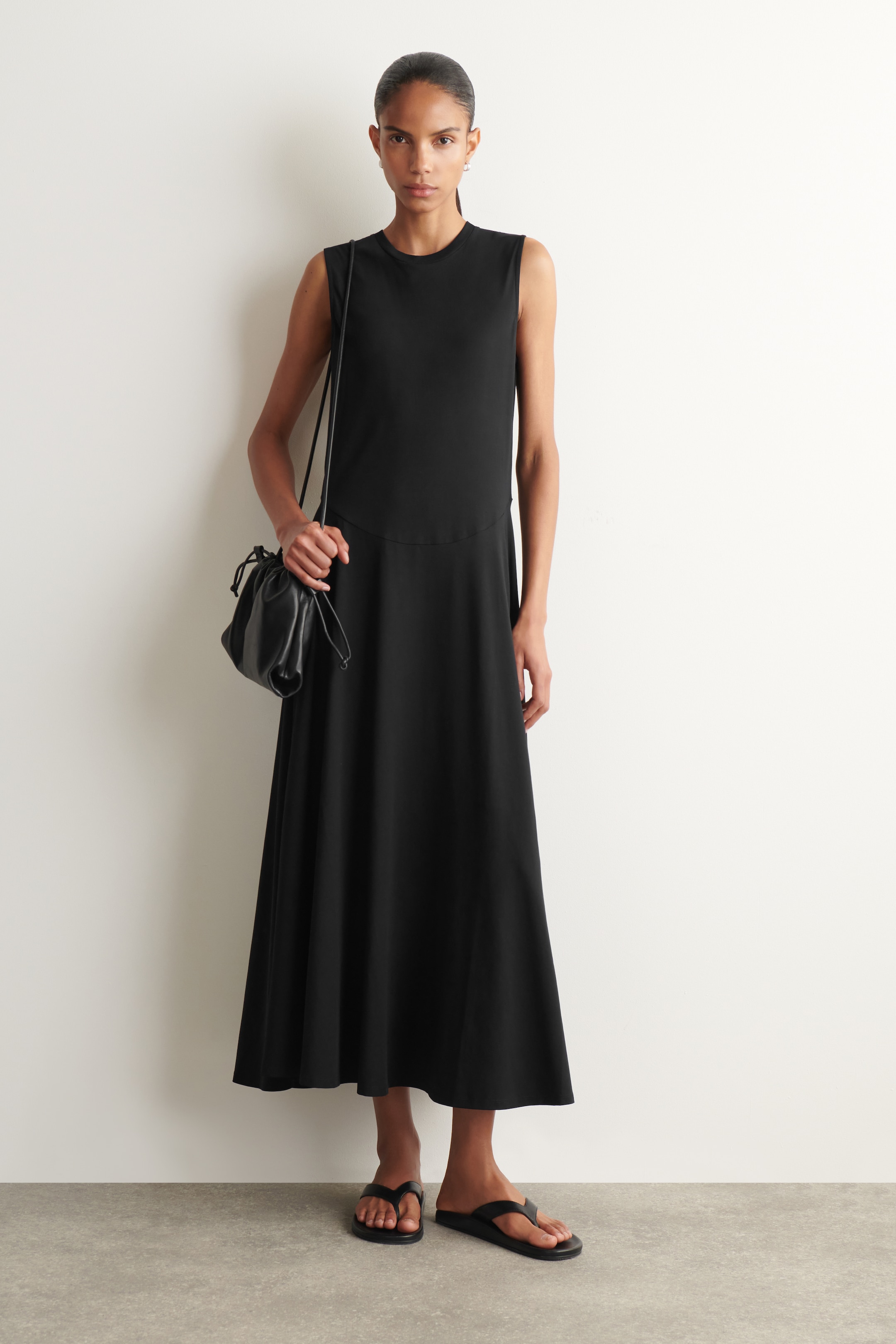 Woman in a sleeveless black dress and sandals, holding a black bag, stands against a plain white wall.