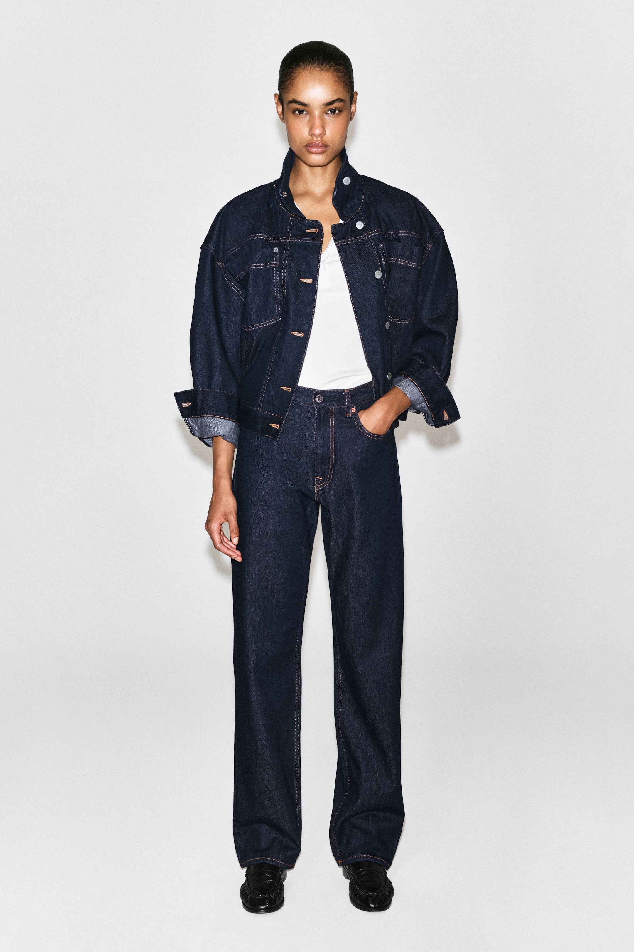 Person wearing a denim jacket and jeans with a white shirt, standing against a plain background.