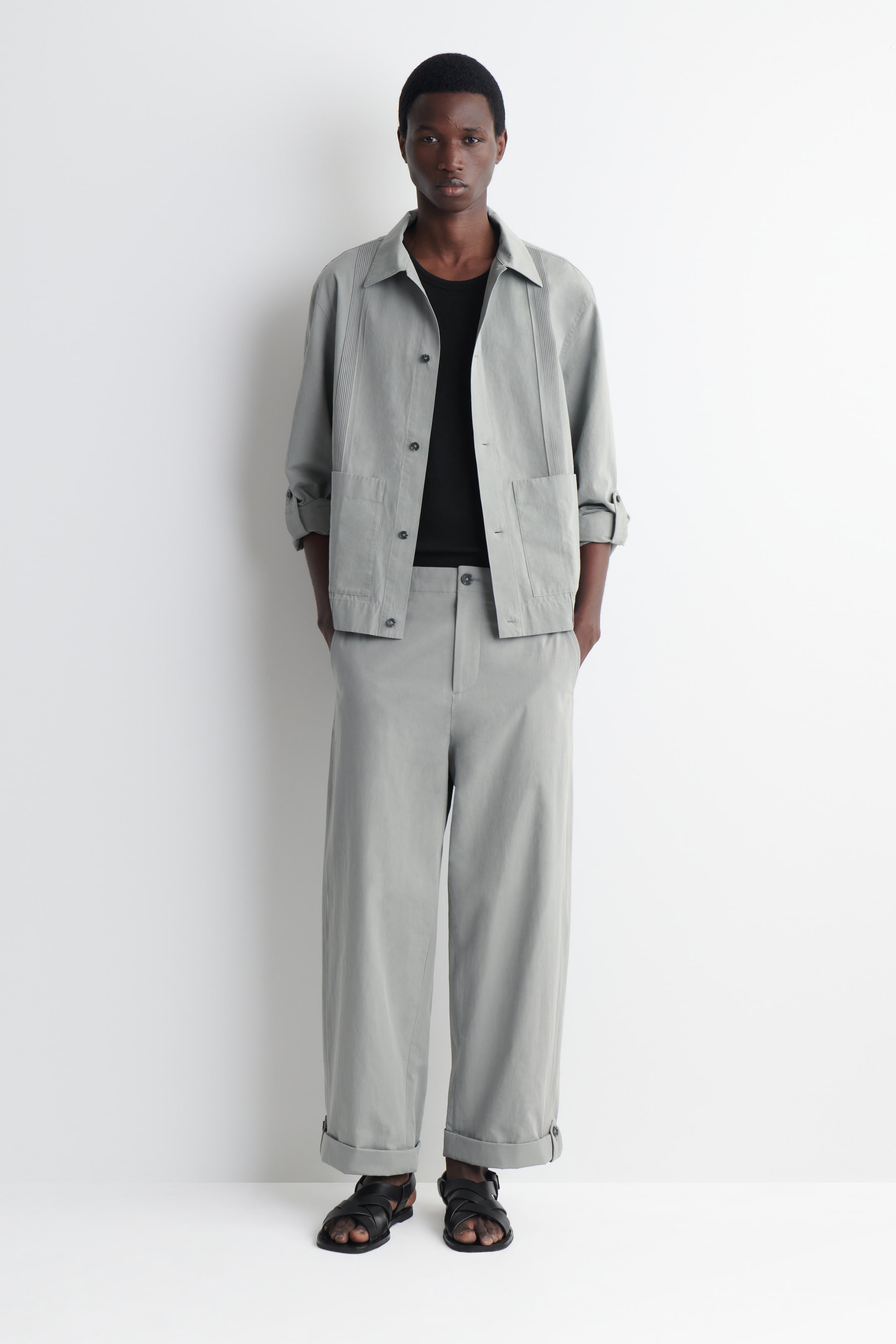 Person dressed in a light gray jacket and pants with a black shirt, standing against a plain white background, wearing black sandals.