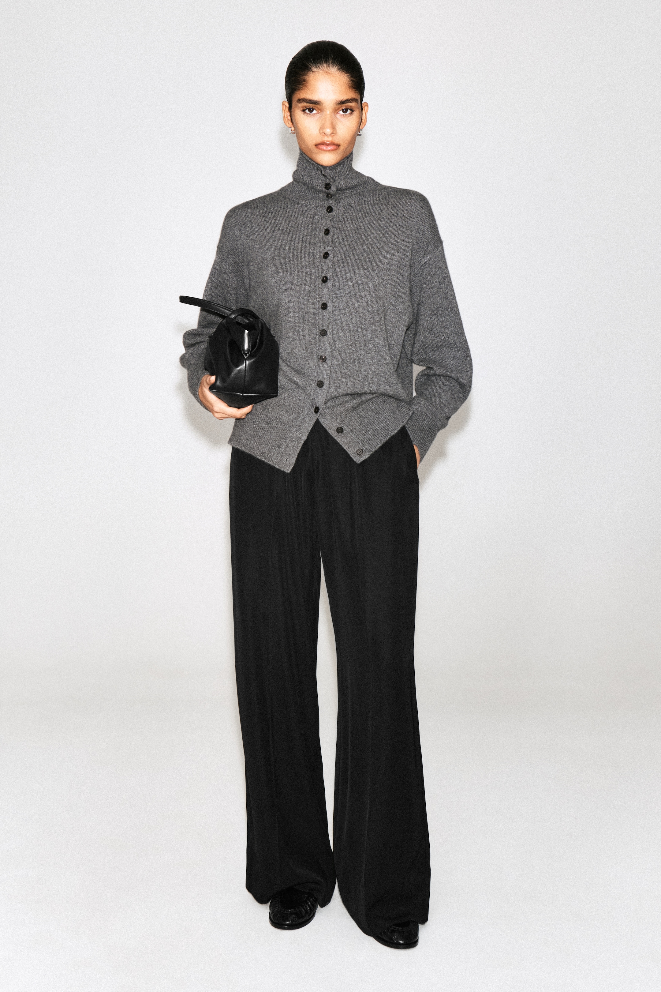 Model in a grey button-up sweater and black wide-leg trousers, holding a small black handbag, against a plain background.