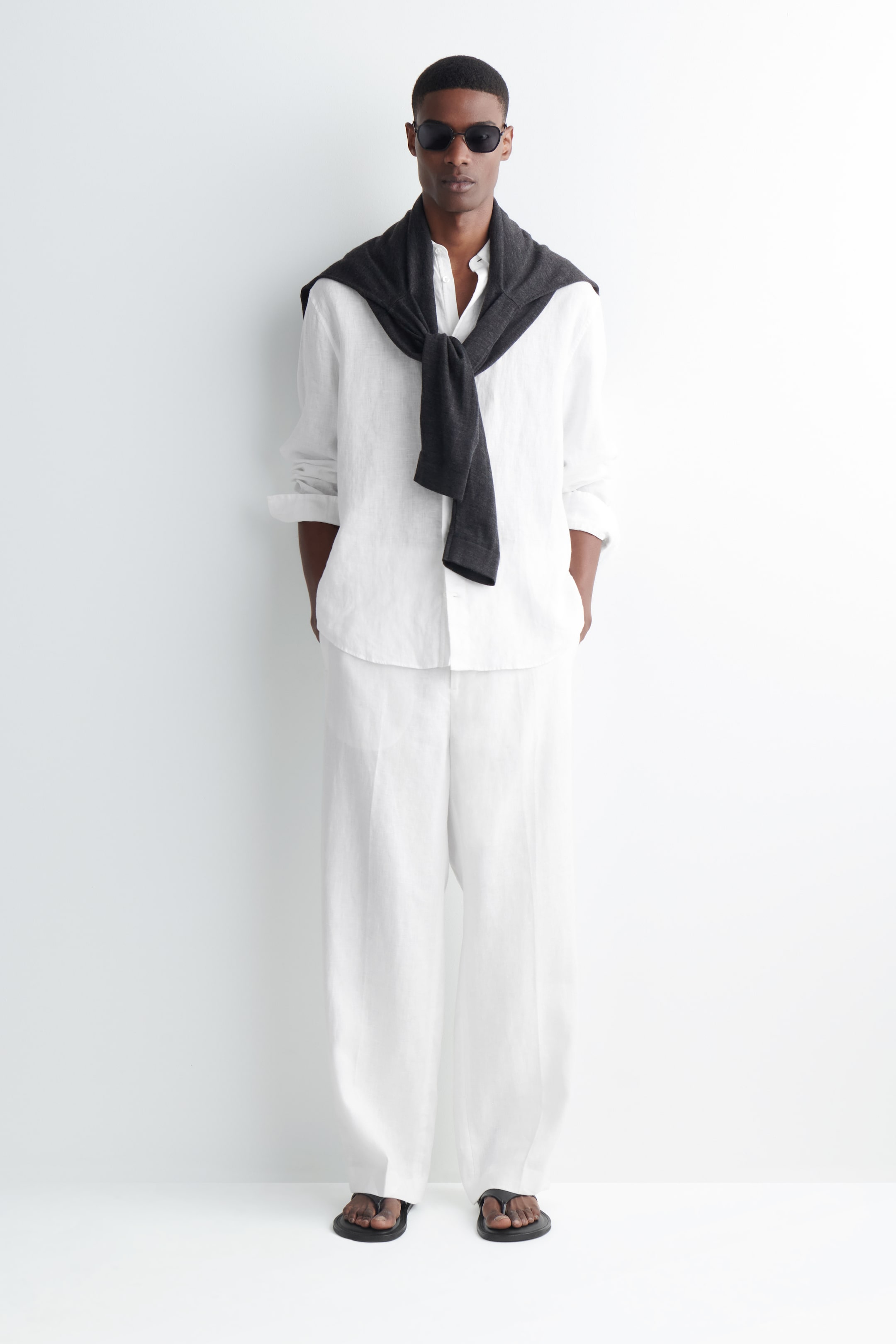 Man in sunglasses wears a white outfit with a sweater over shoulders, standing against a white background, hands in pockets, wearing sandals.