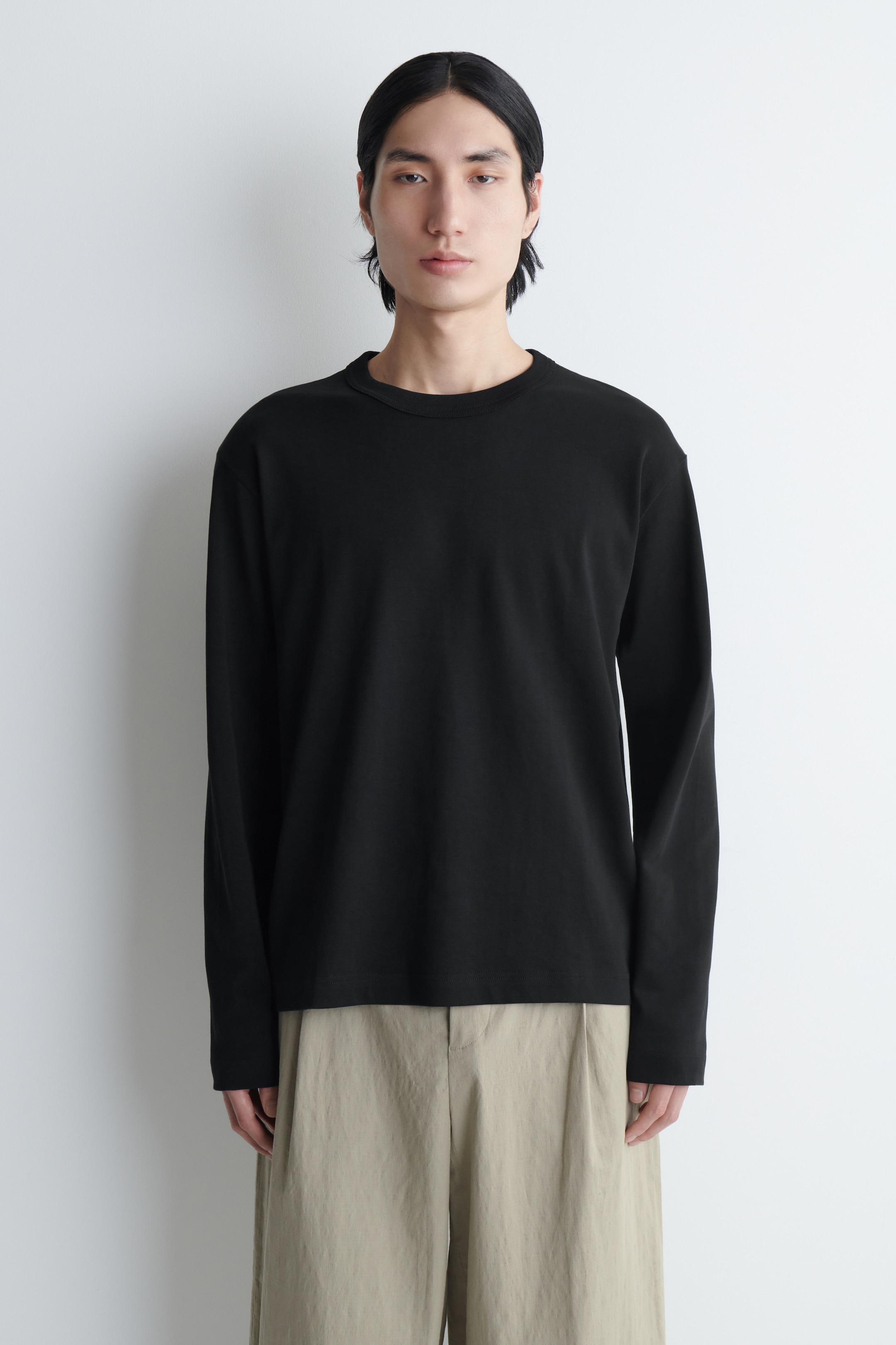 A person with long black hair wearing a black long-sleeved shirt and beige pants, standing against a white background.