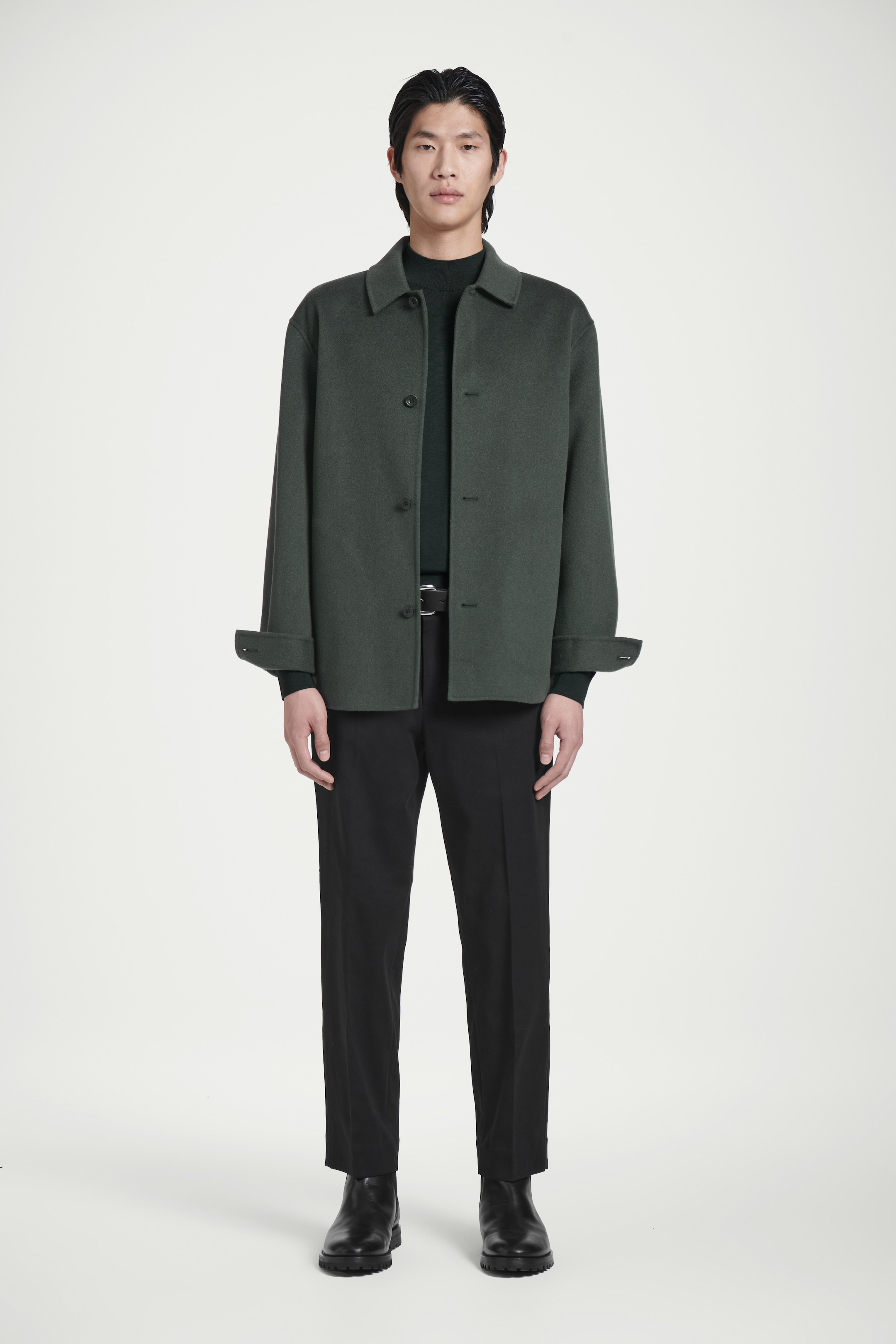 A person stands wearing a dark green jacket, black pants, and black boots against a plain white background.