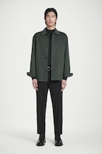 Placeholder for: A person stands wearing a dark green jacket, black pants, and black boots against a plain white background.