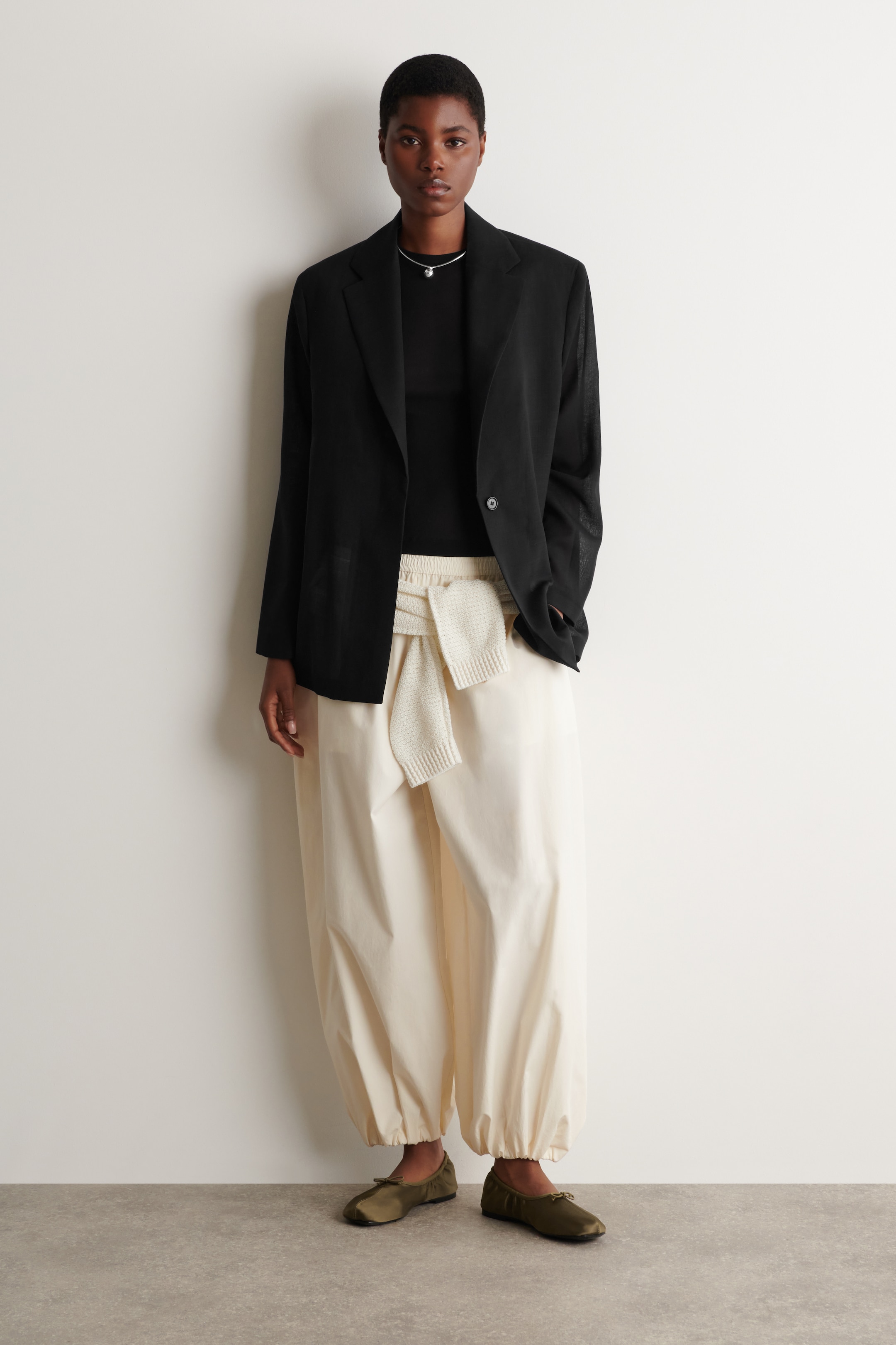 Person in a black blazer, black shirt, beige pants with a tied sweater, and olive-green shoes, standing against a white wall.