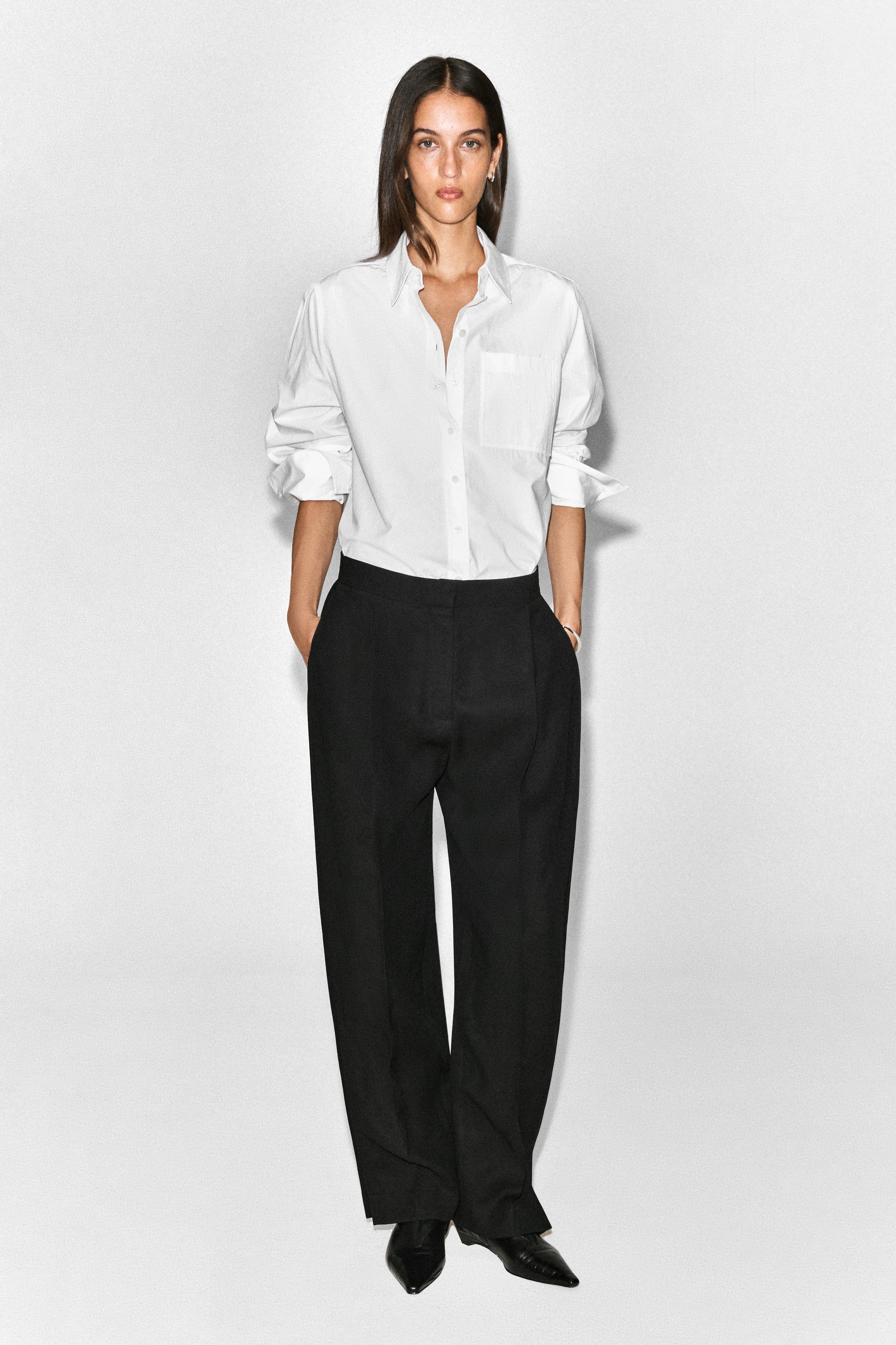 A woman stands against a plain white background, wearing a white button-up shirt and black wide-leg trousers, with hands in pockets.