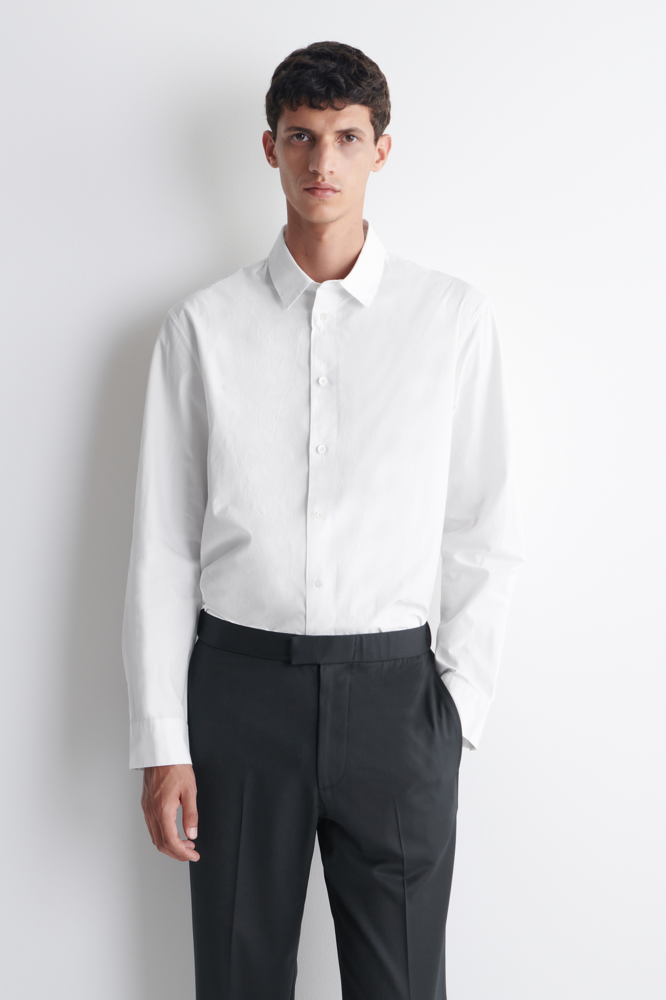 A person wearing a white dress shirt and black trousers, standing against a white background.
