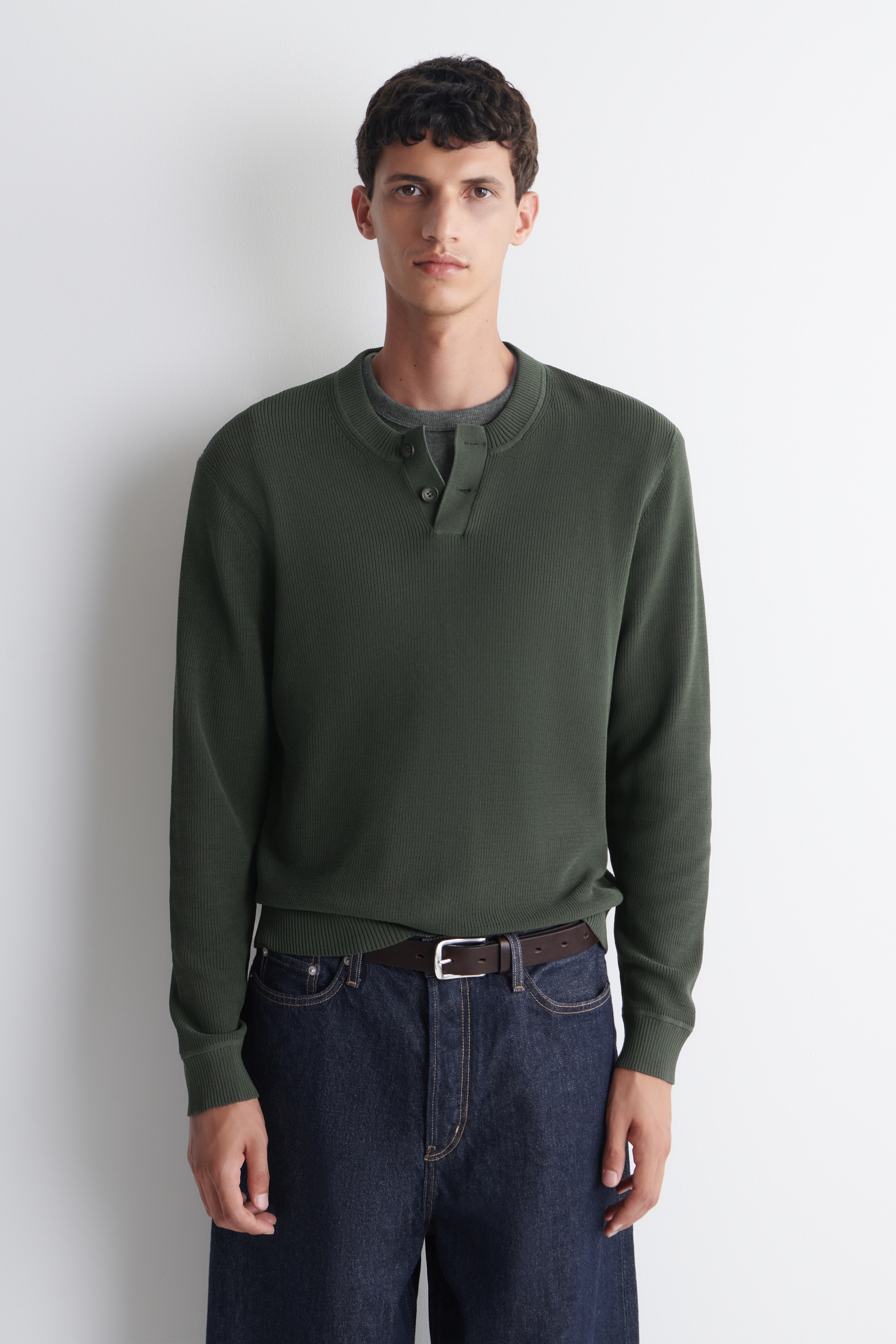 A person wearing a dark green henley sweater and blue jeans, standing against a plain white background.