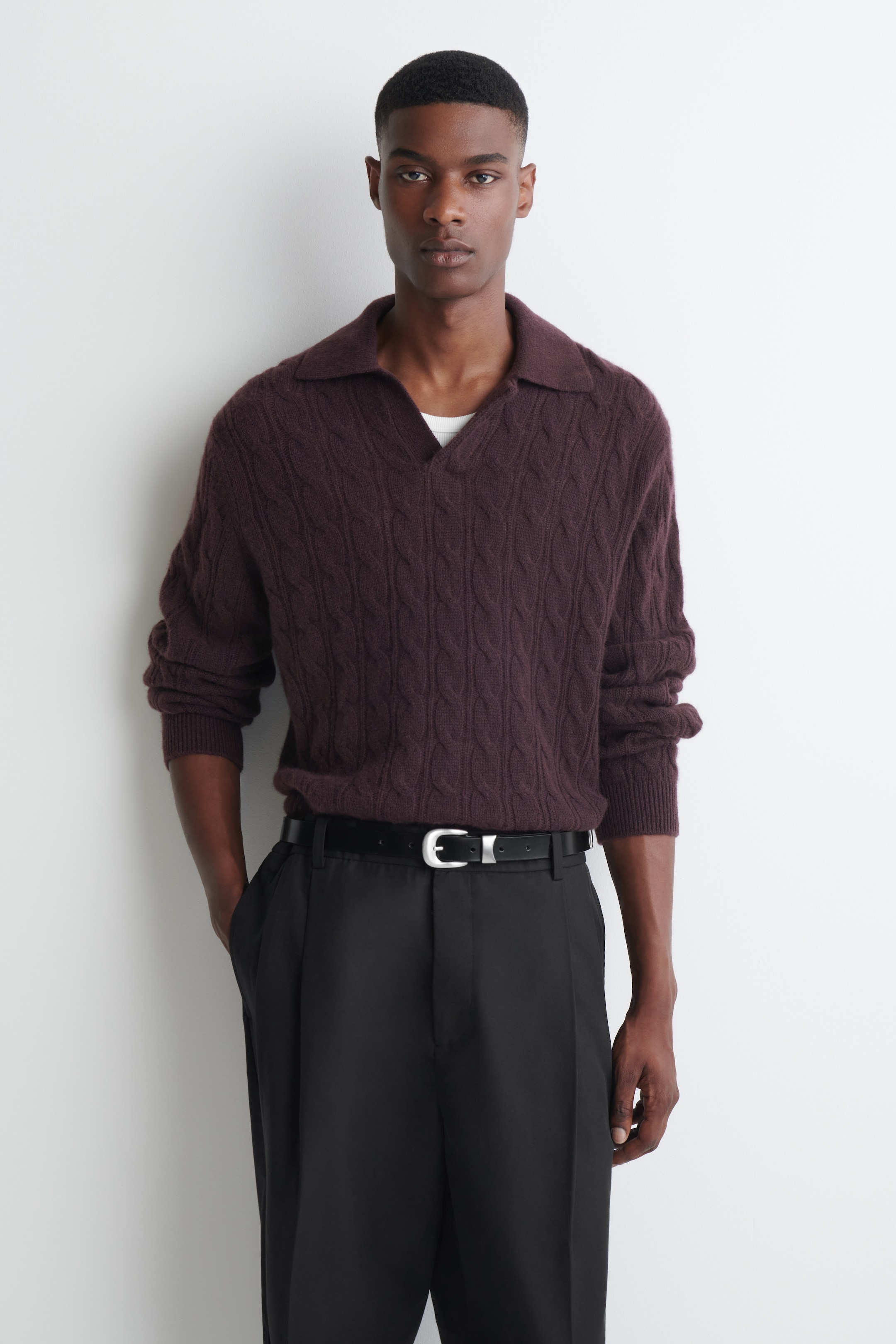 A man wearing a maroon cable-knit sweater and black trousers stands against a white background.