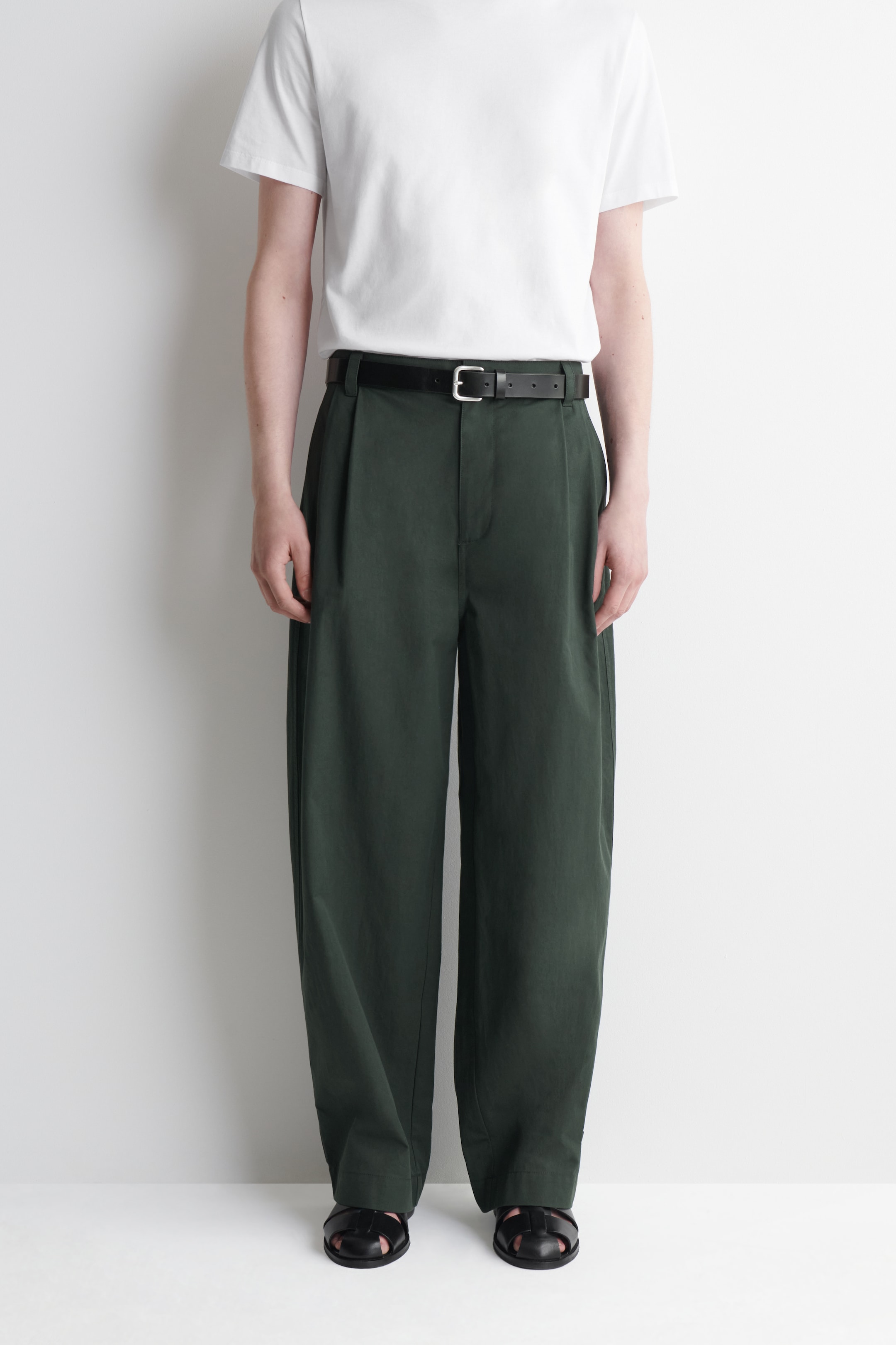 Person wearing a white T-shirt, dark green high-waisted trousers with a black belt, and black sandals, standing against a white background.