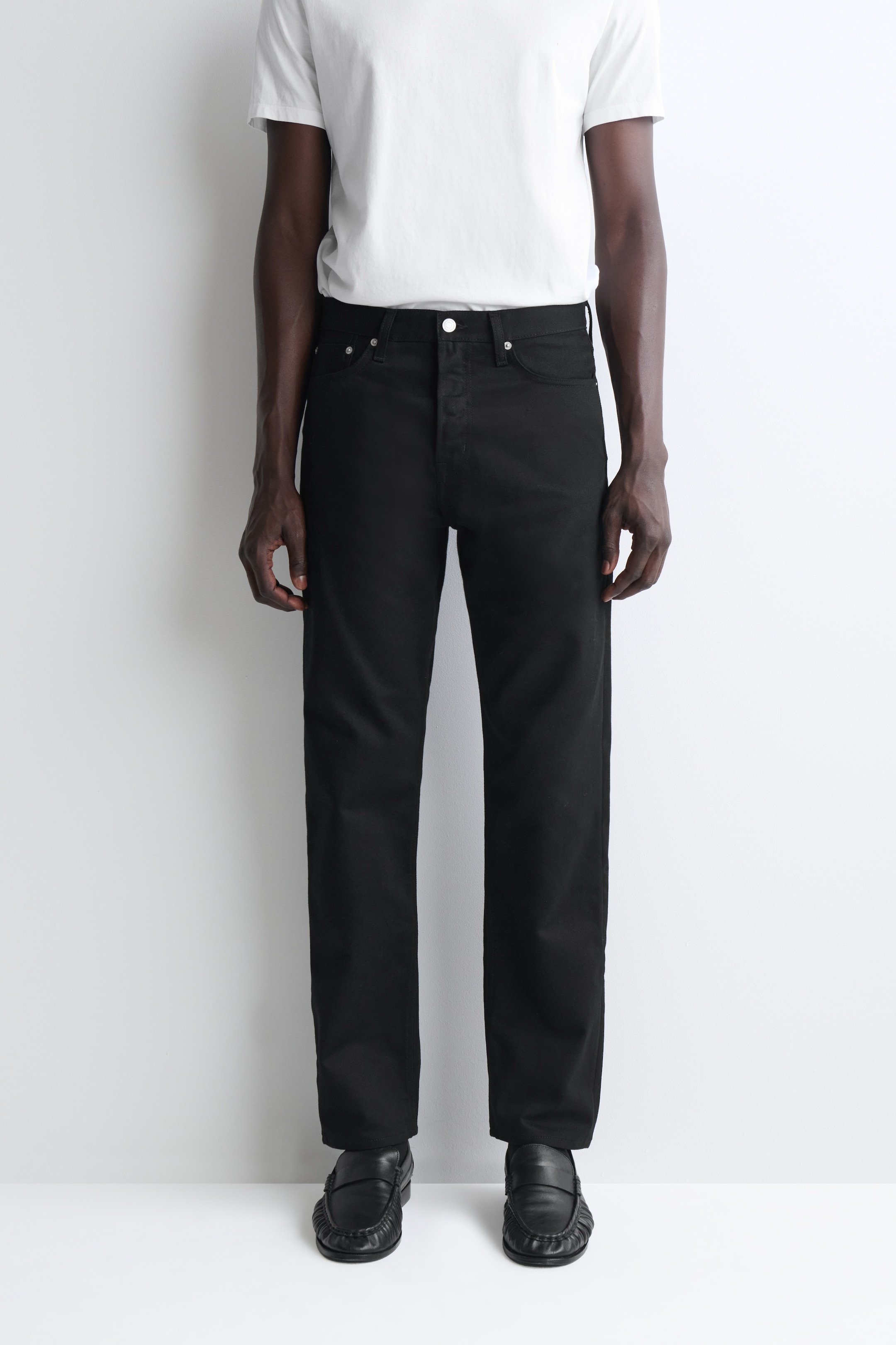 A person wearing a white T-shirt, black pants, and black shoes standing against a plain white background.